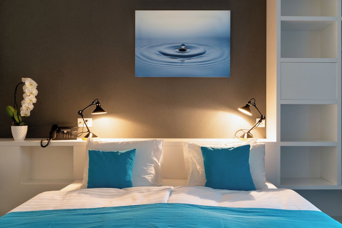 Habitación de hotel con una cama, dos lámparas de noche, un teléfono y un cuadro colgado en la pared con forma de gota de agua.