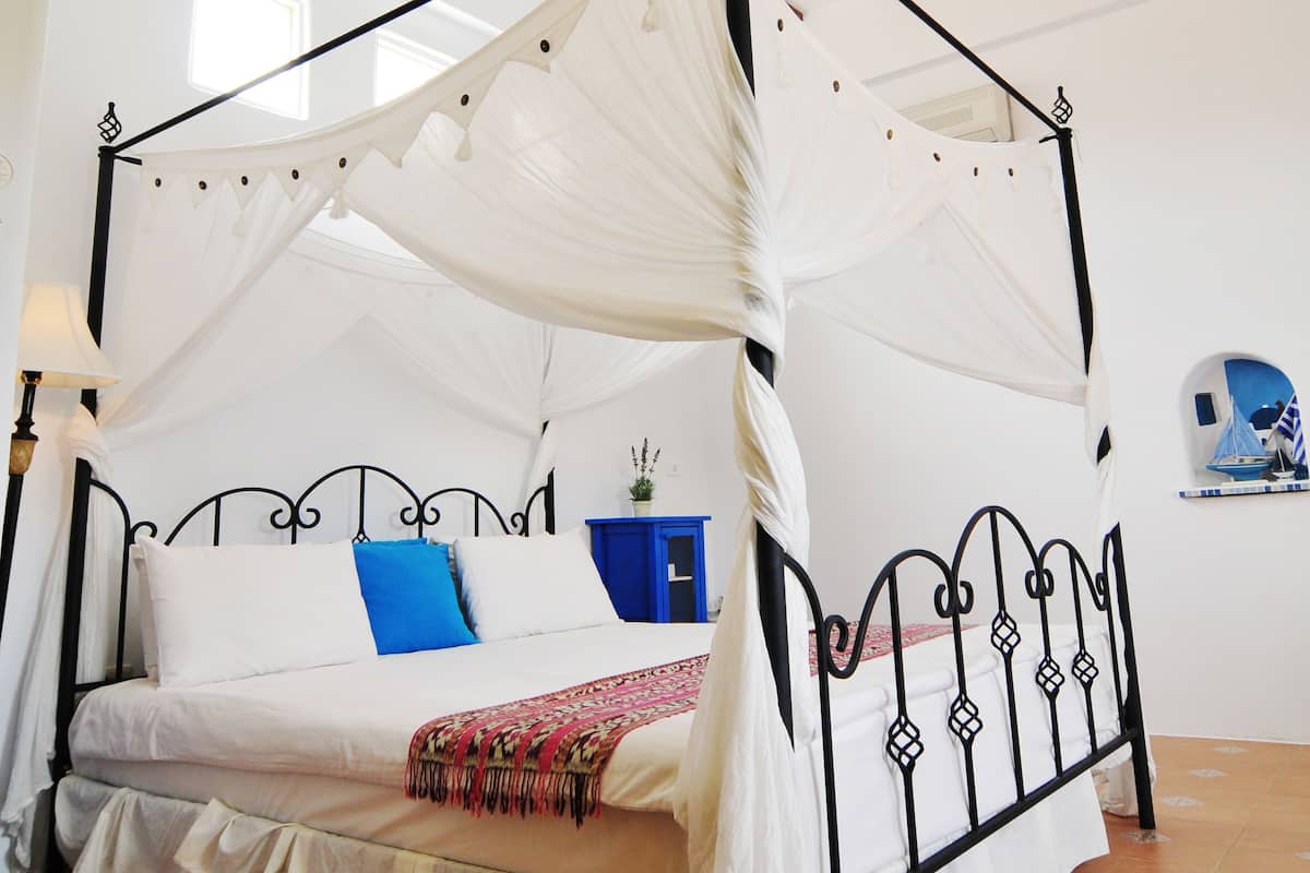 Una cama con dosel y cuatro columnas, con ropa de cama blanca y una almohada azul, ubicada en una habitación con baldosas de barro cocido y vigas de madera en el techo.