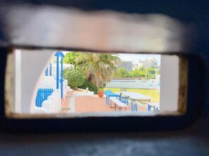 A view through a window with a blue railing, a palm tree, and a building in the background. - Greek frontier villa (Magong)