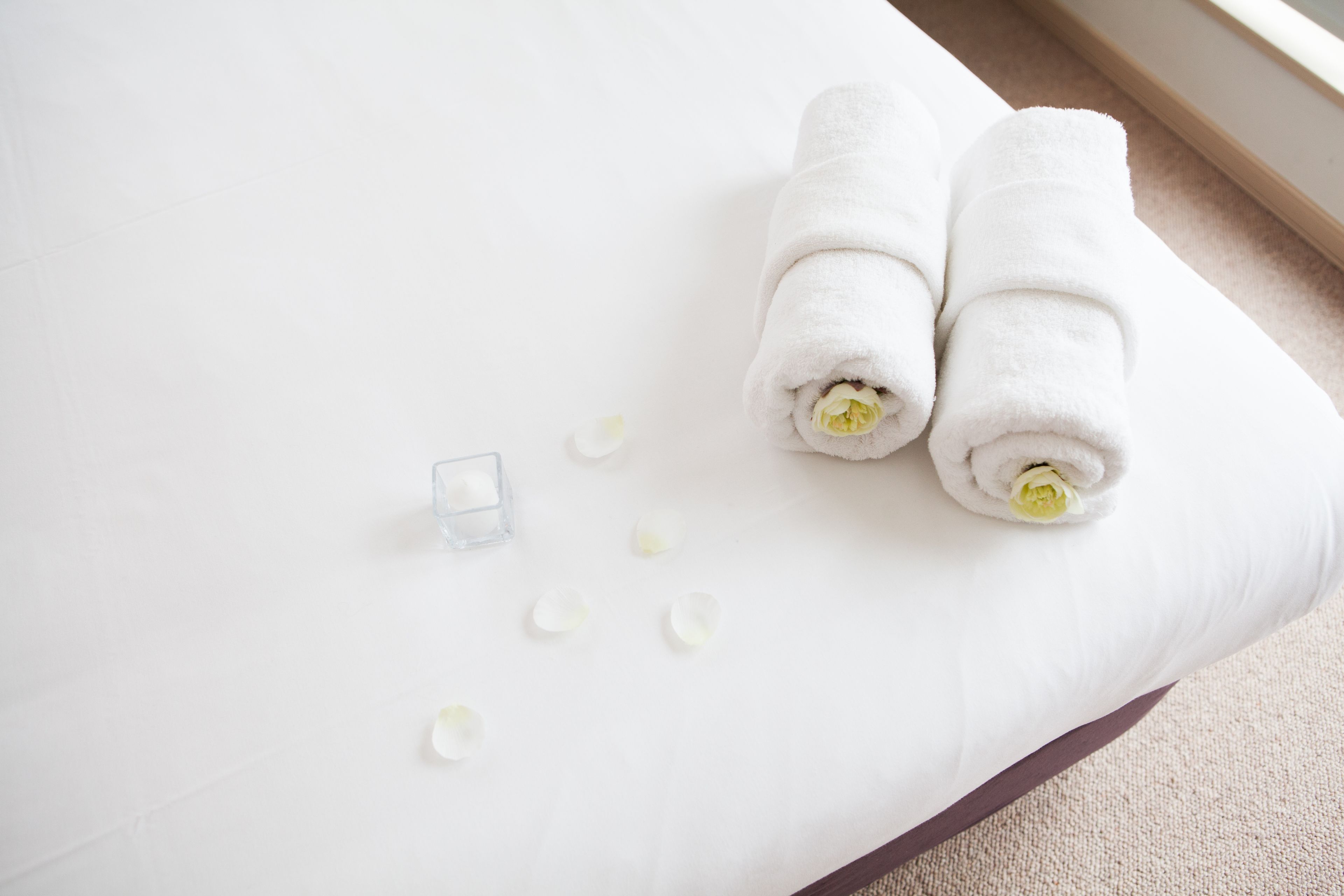 A white bed with two rolled towels and a glass of water on a white mattress.