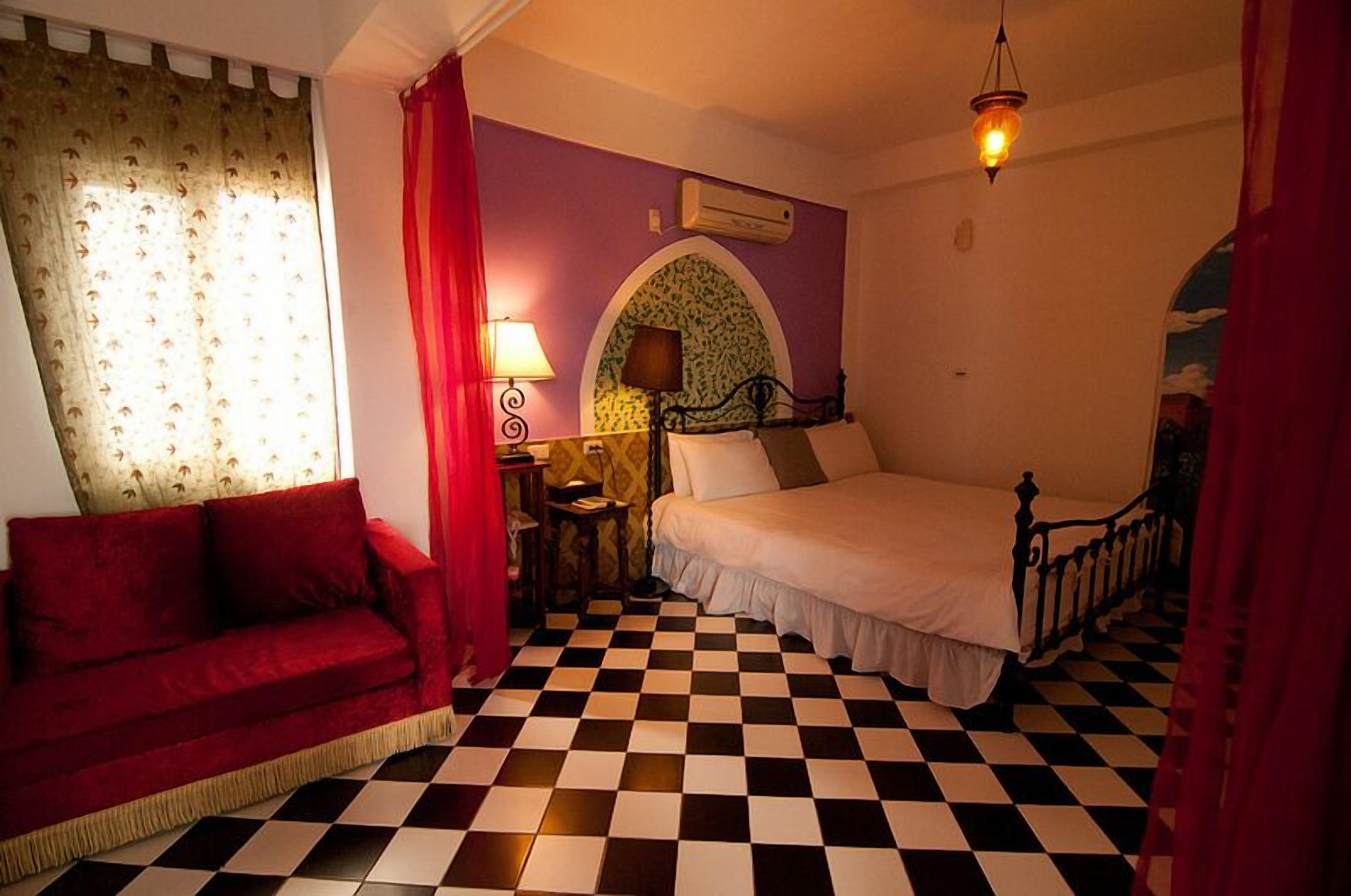 A room with a checkered floor, a bed with a white bedspread, a red sofa, and a lamp on a nightstand.