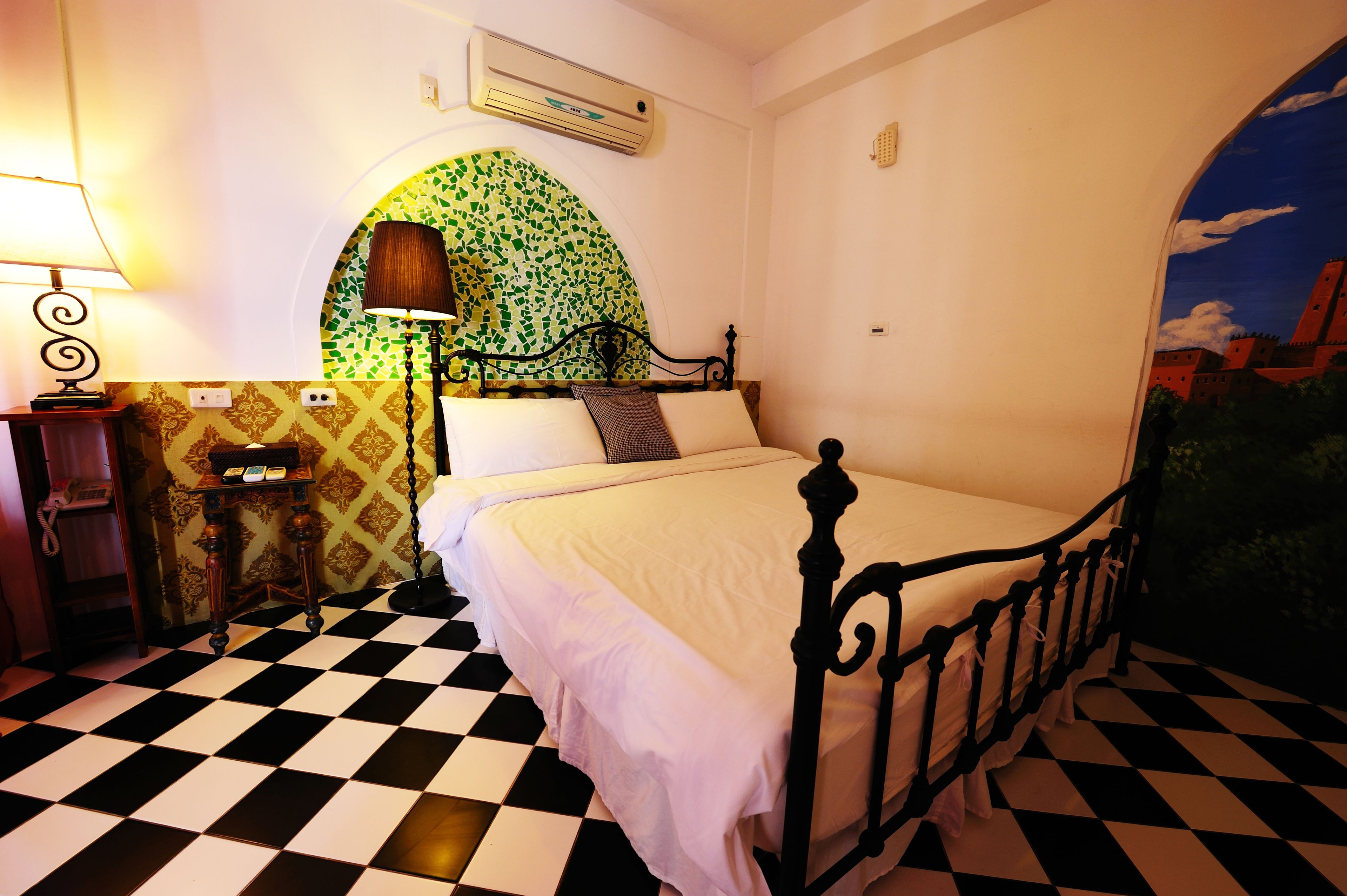 A room with a checkered floor, a bed with a metal frame, a lamp, a wall-mounted air conditioner, and a decorative archway with a green mosaic pattern.