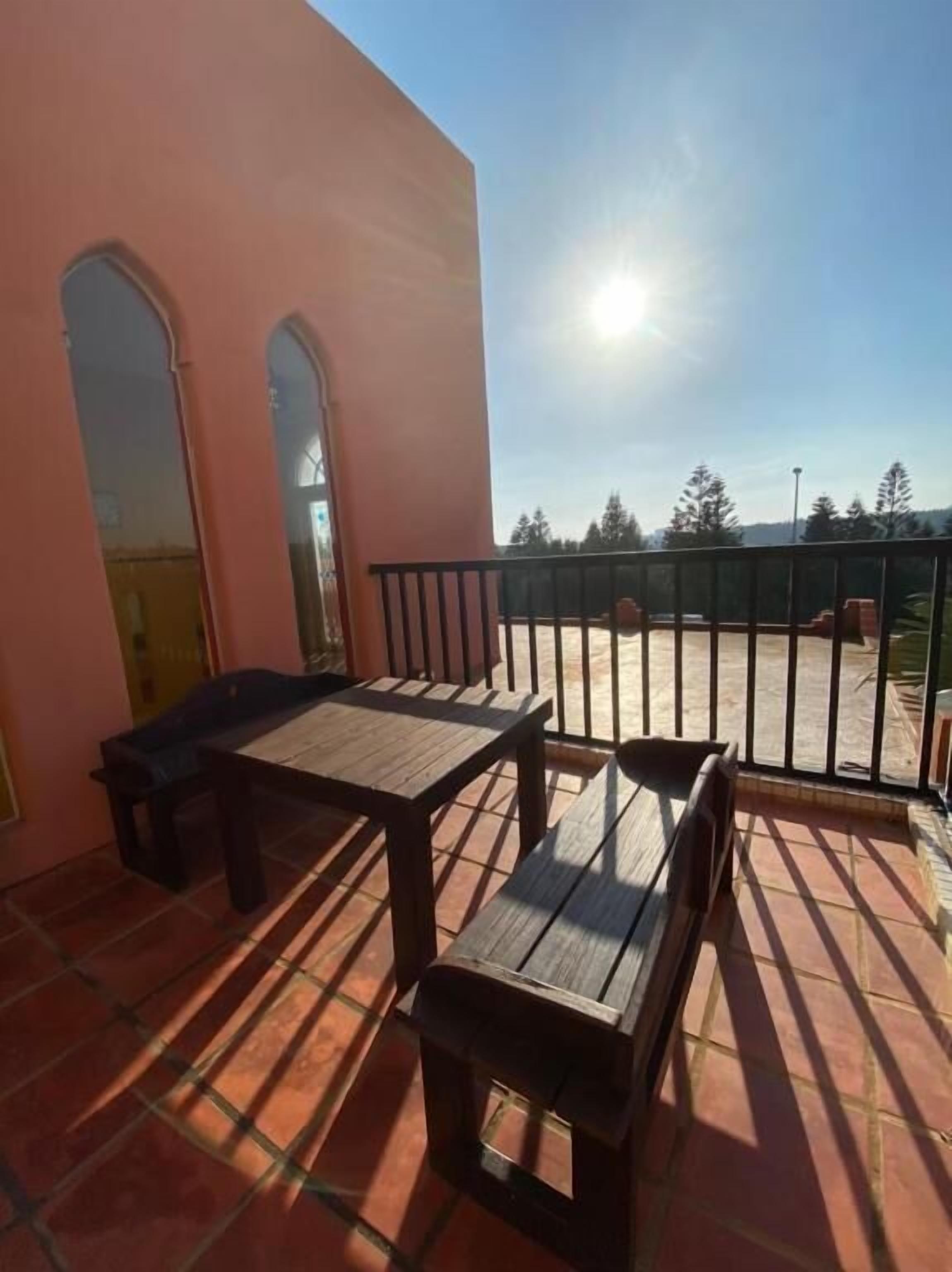 Uma varanda com mesa e cadeiras, guarda-corpo de metal, e vista para árvores e um edifício ao fundo.