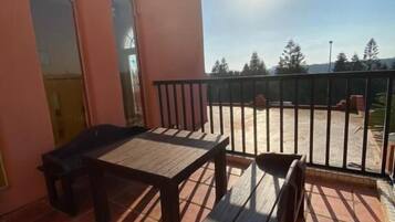 Uma varanda com mesa e cadeiras, guarda-corpo de metal, e vista para árvores e um edifício ao fundo.