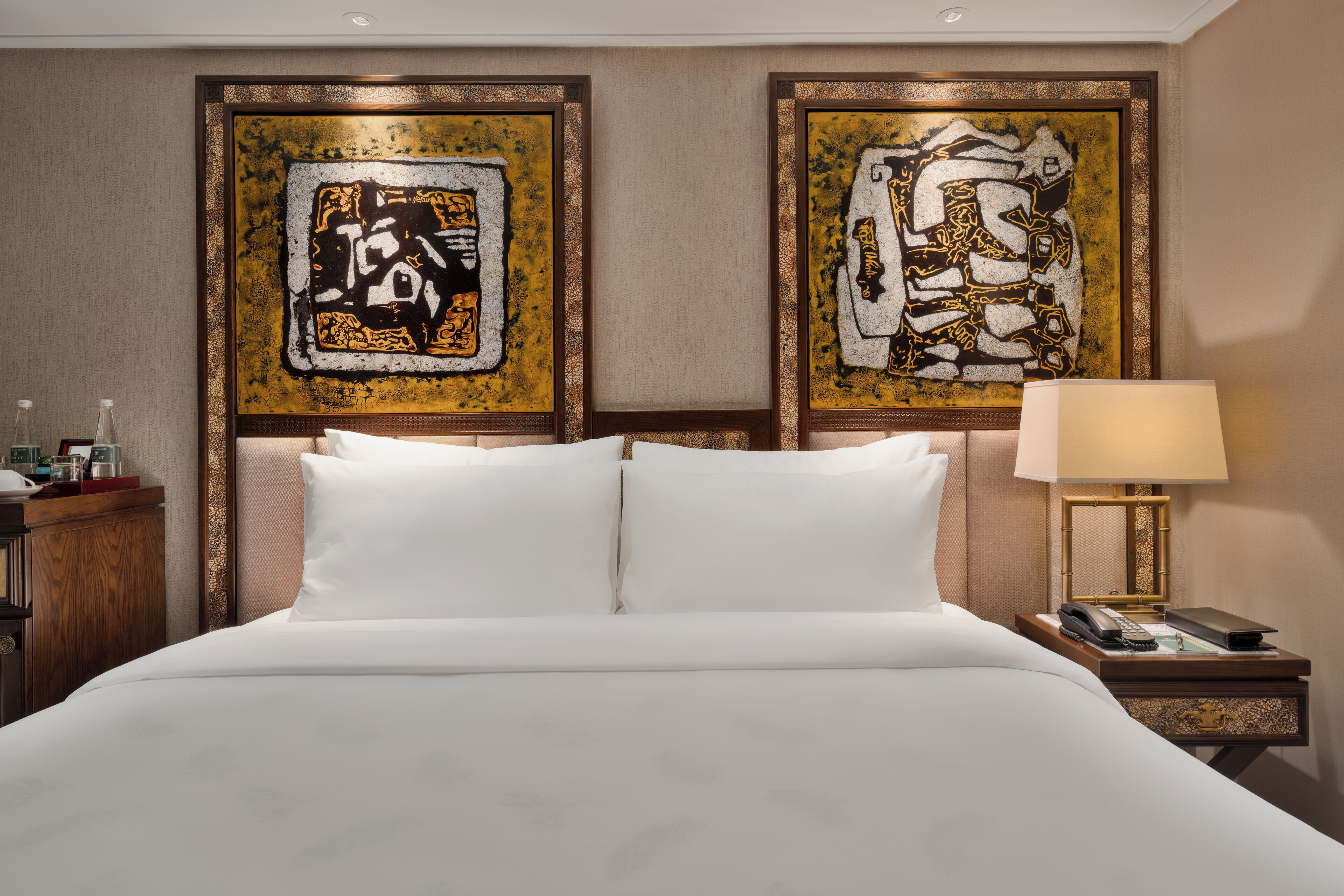 A hotel room with a large bed, two framed abstract paintings on the wall, a nightstand with a lamp, and a small table with a phone and remote control.