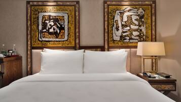 A hotel room with a large bed, two framed abstract paintings on the wall, a nightstand with a lamp, and a small table with a phone and remote control.