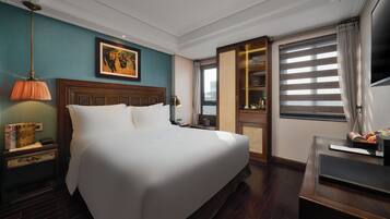 A hotel room with a large bed, a nightstand with a lamp, a television, and a window with blinds.