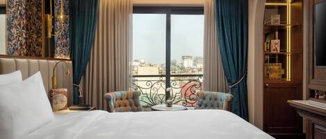 A hotel room with a large bed, two armchairs, a small table, and a view of the city through a large window.