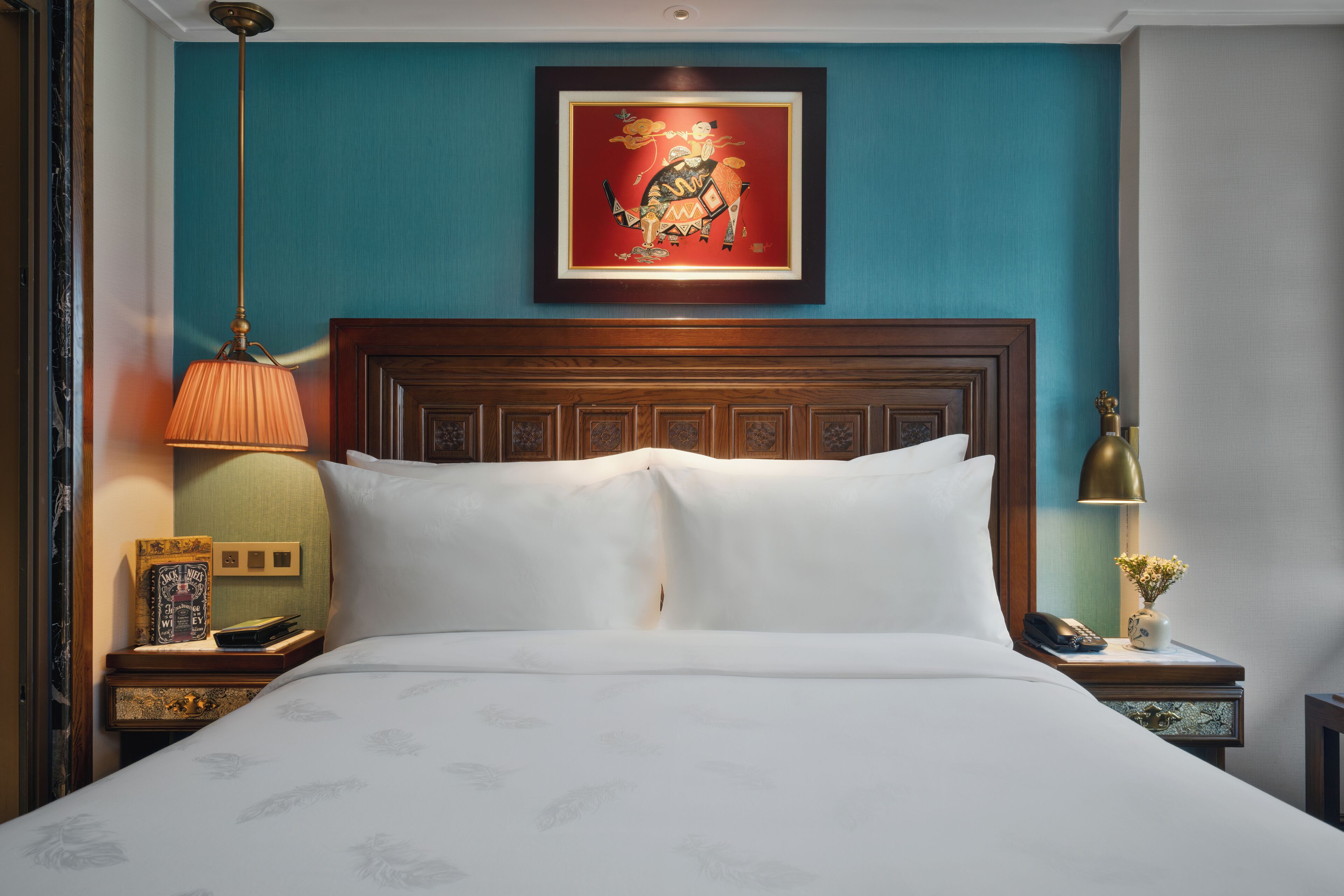 A neatly made bed with a headboard, a framed artwork above it, and a nightstand with a lamp and a vase of flowers.