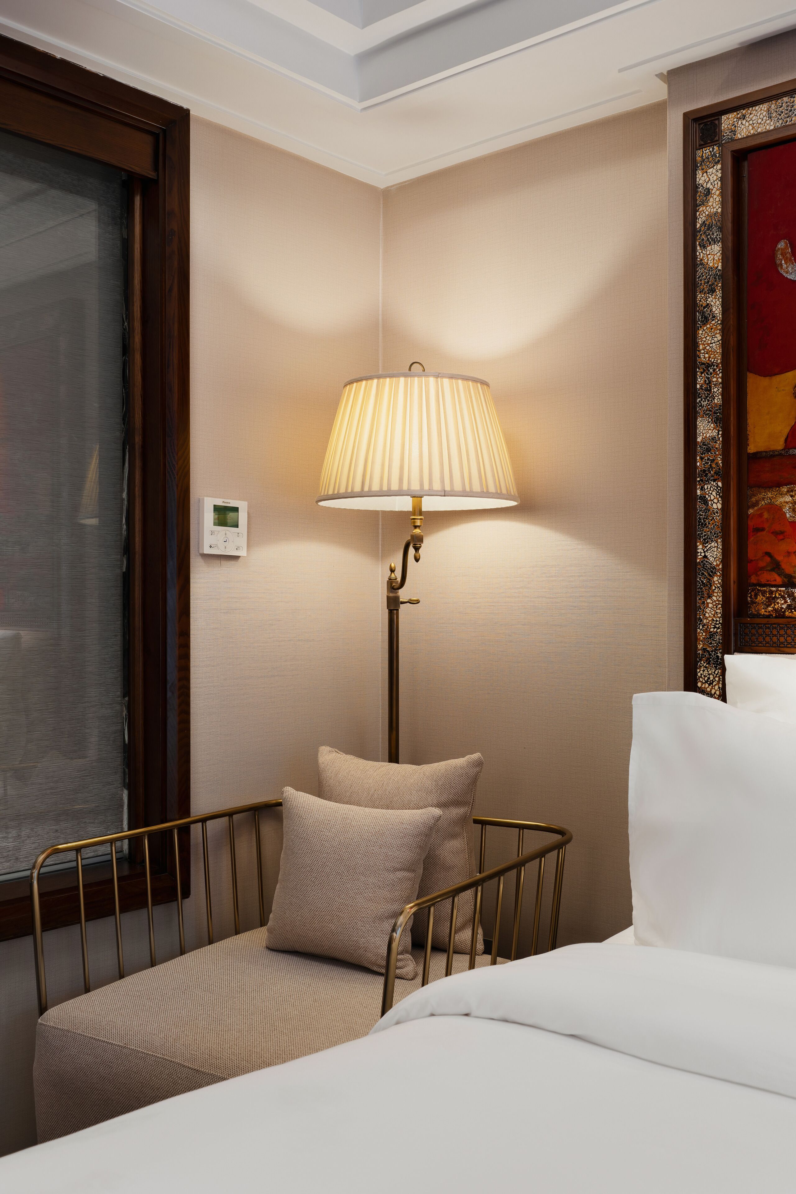 A hotel room with a bed, a bedside lamp, a thermostat, and a window with a curtain.