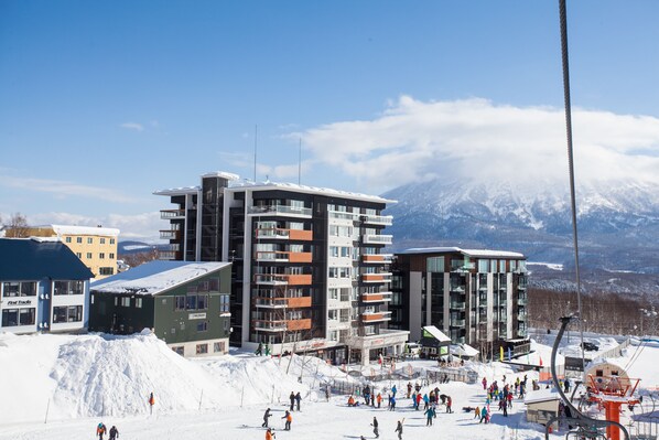 A modern ski resort with multi-story buildings, a ski lift, and a snowy mountain backdrop. - Alpen Ridge (Kutchan)