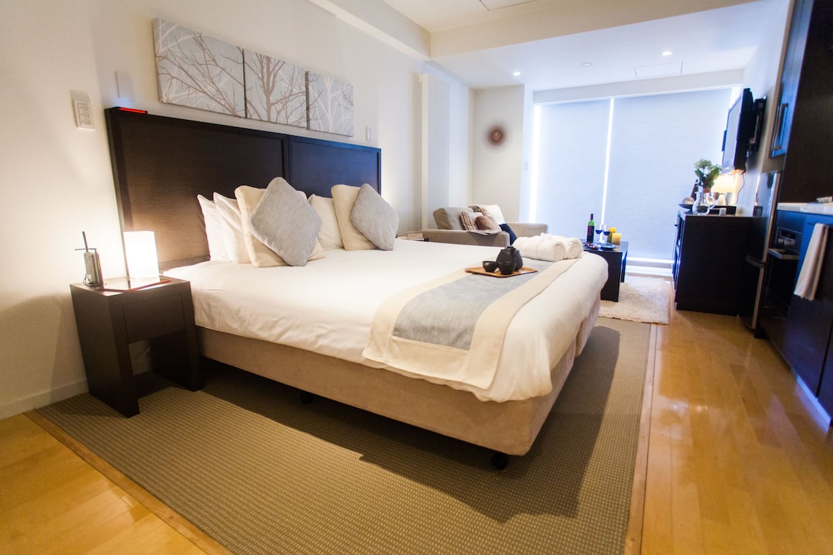 A modern hotel room with a large bed, a bedside table with a lamp, a small seating area with a sofa, and a TV mounted on the wall.