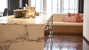 A kitchen with a marble countertop, a stove, and a sink. There is a sofa with a patterned cushion in the background.