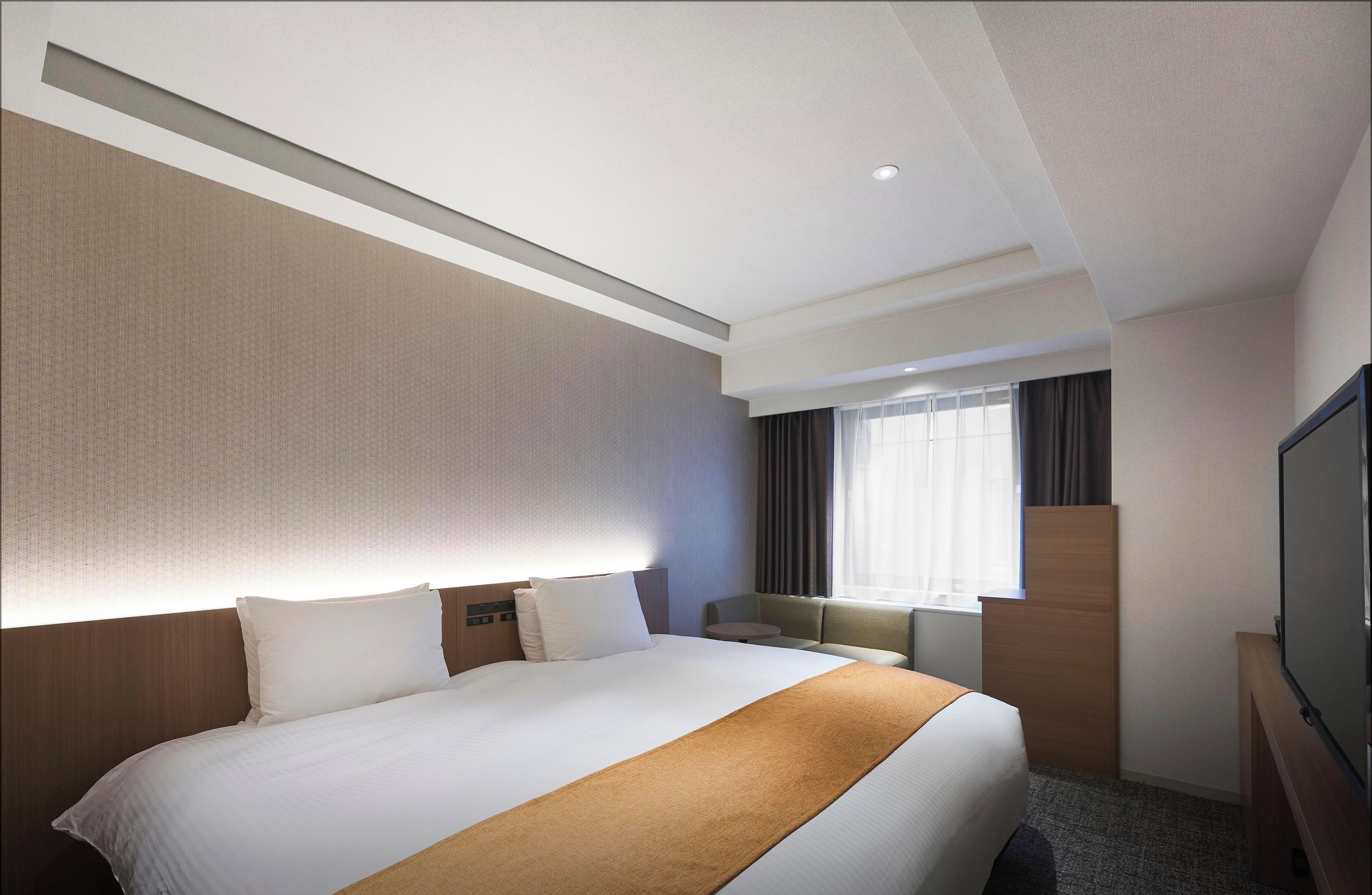 Ein Hotelzimmer mit einem großen Bett, einem Flachbildfernseher, einem Fenster mit Vorhängen und einem kleinen Tisch.
