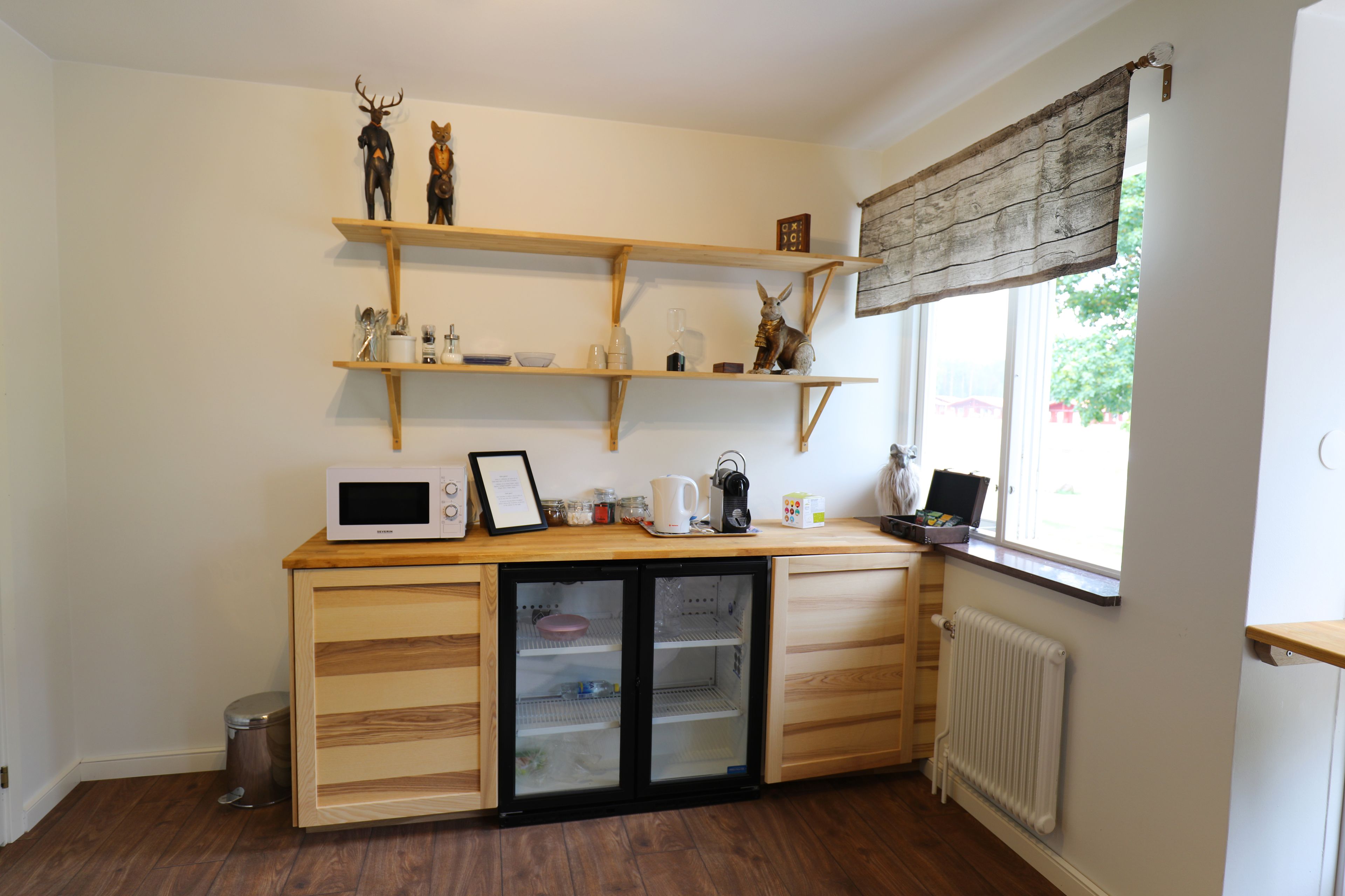Una cocina compacta con armarios de madera, microondas integrado y un refrigerador pequeño. Estantes superiores exhiben diversos utensilios y adornos de cocina.