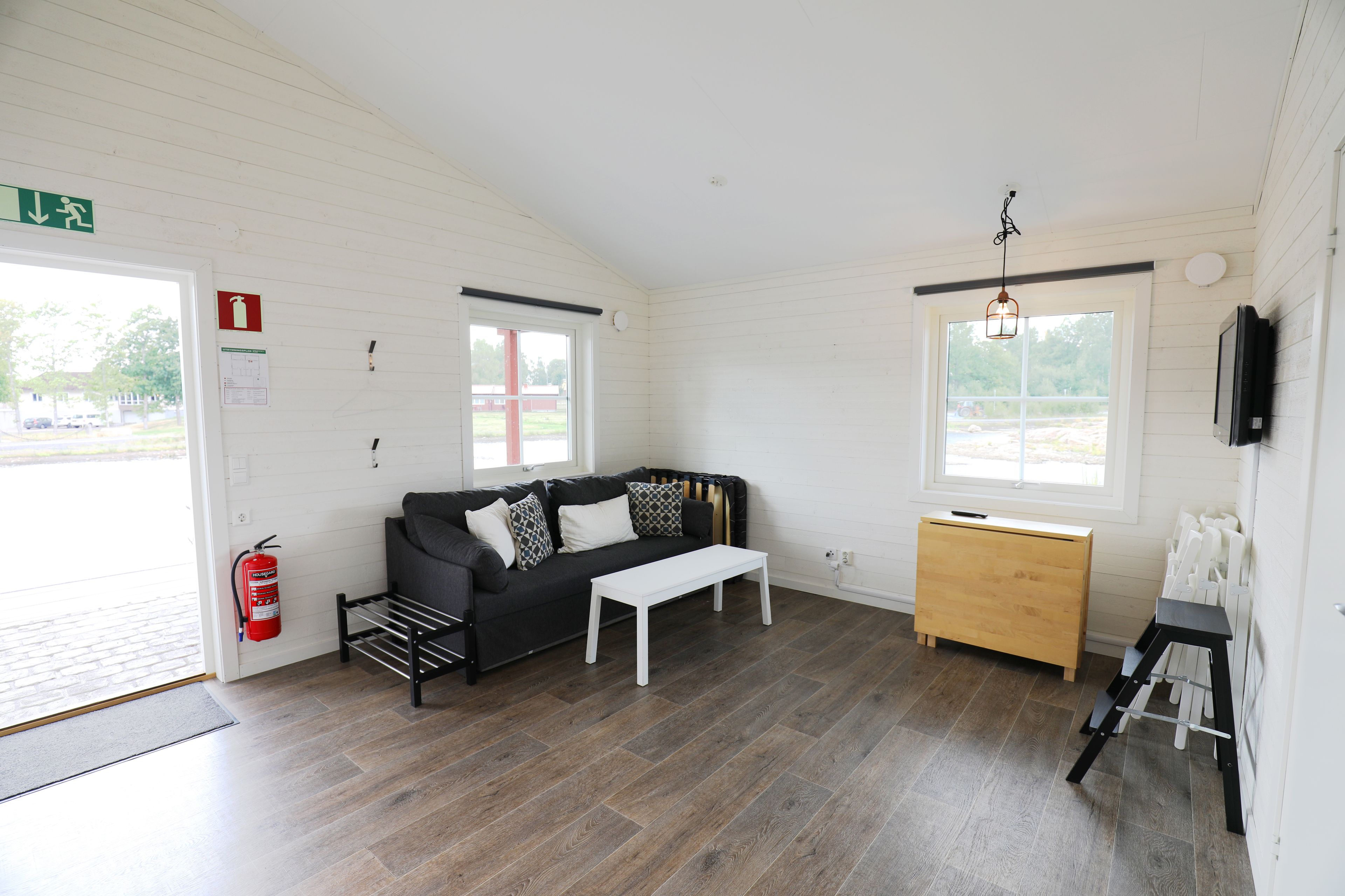Habitación con sofá negro, mesa de centro blanca y un mueble de madera. Hay un extintor de incendios en la pared y una ventana con vistas al exterior.