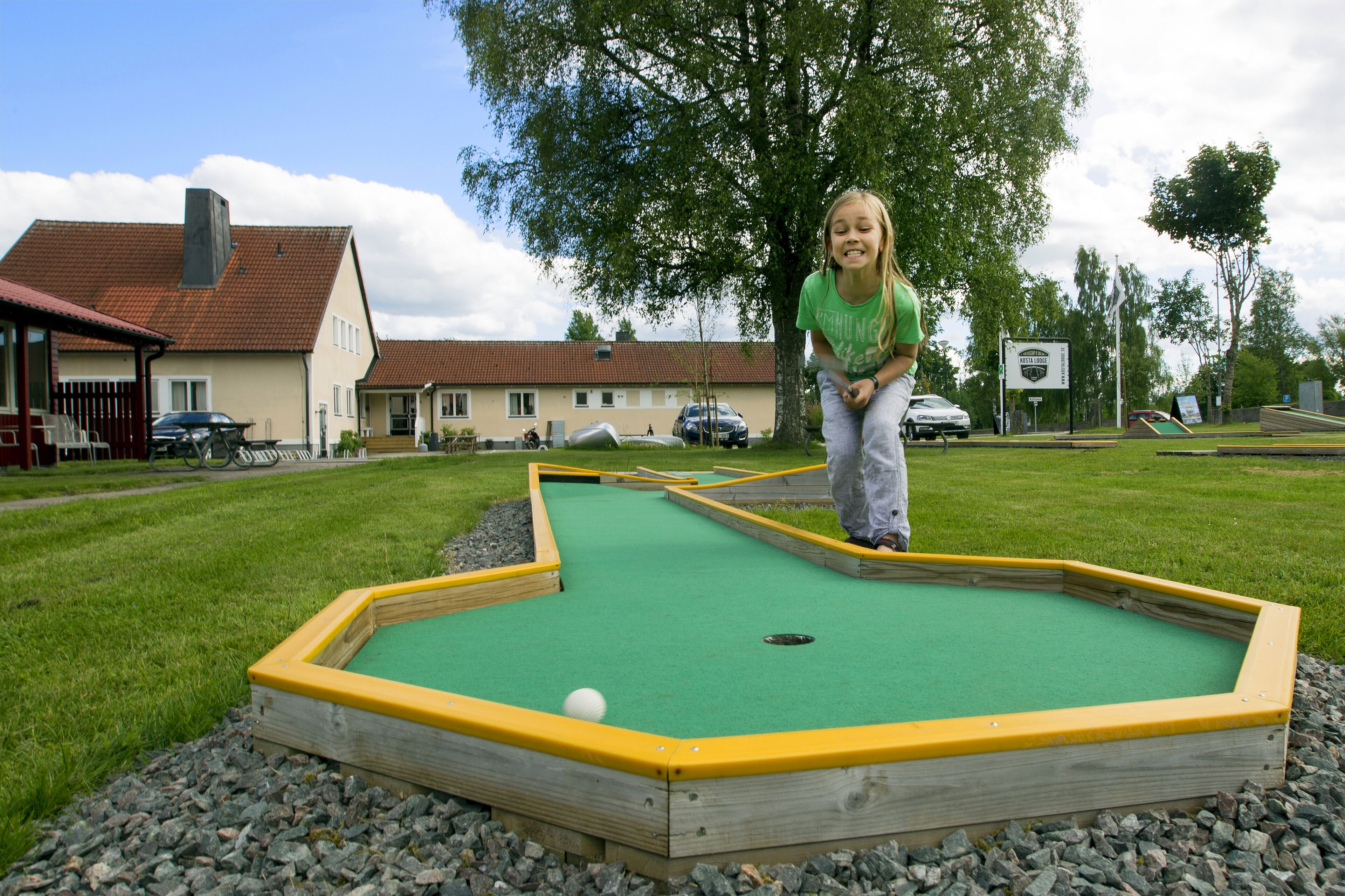 A miniature golf course with a green playing surface and yellow borders, surrounded by a grassy area and a building in the background.