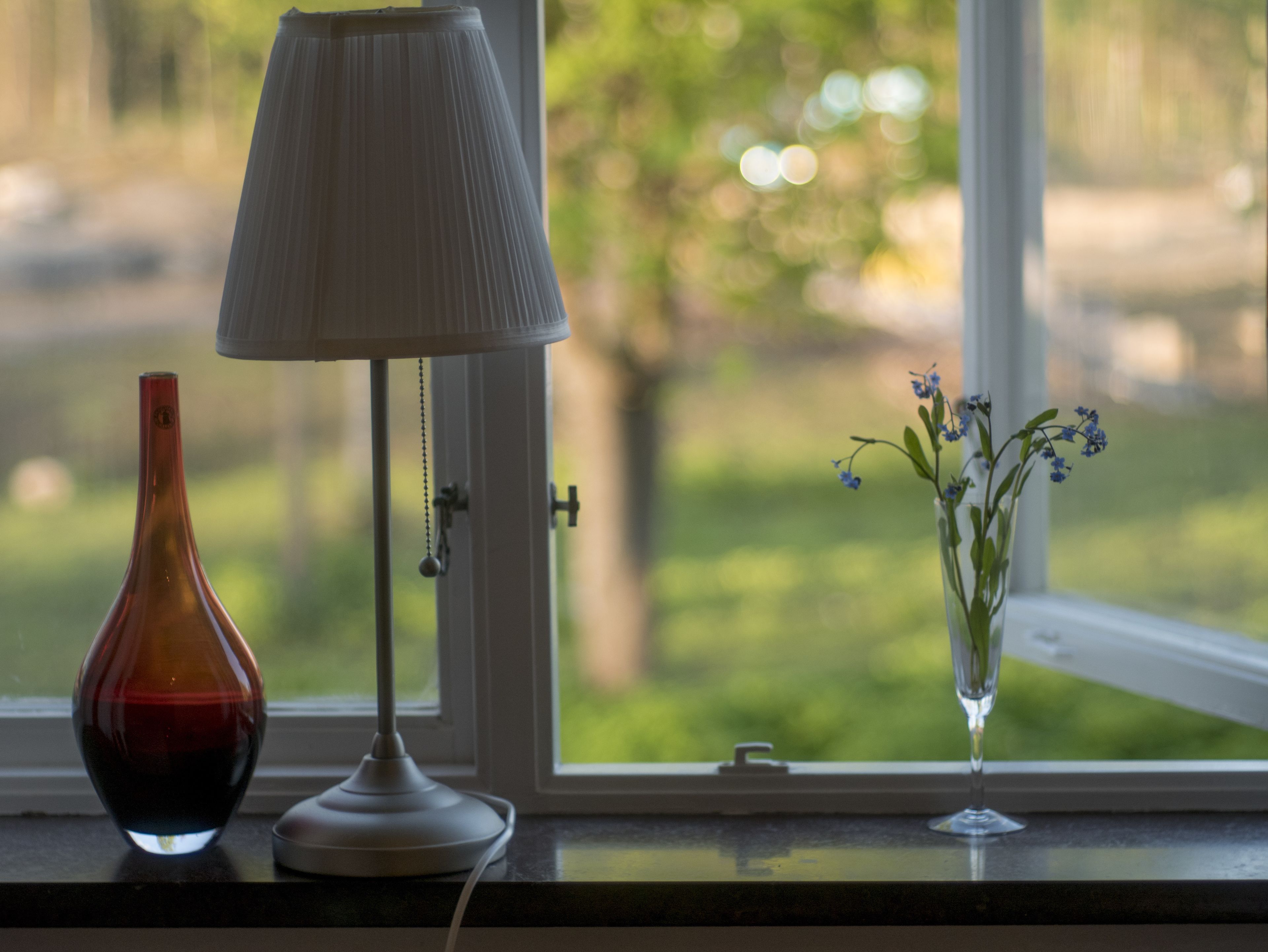 A lamp, a vase with flowers, and a window with a view of greenery.