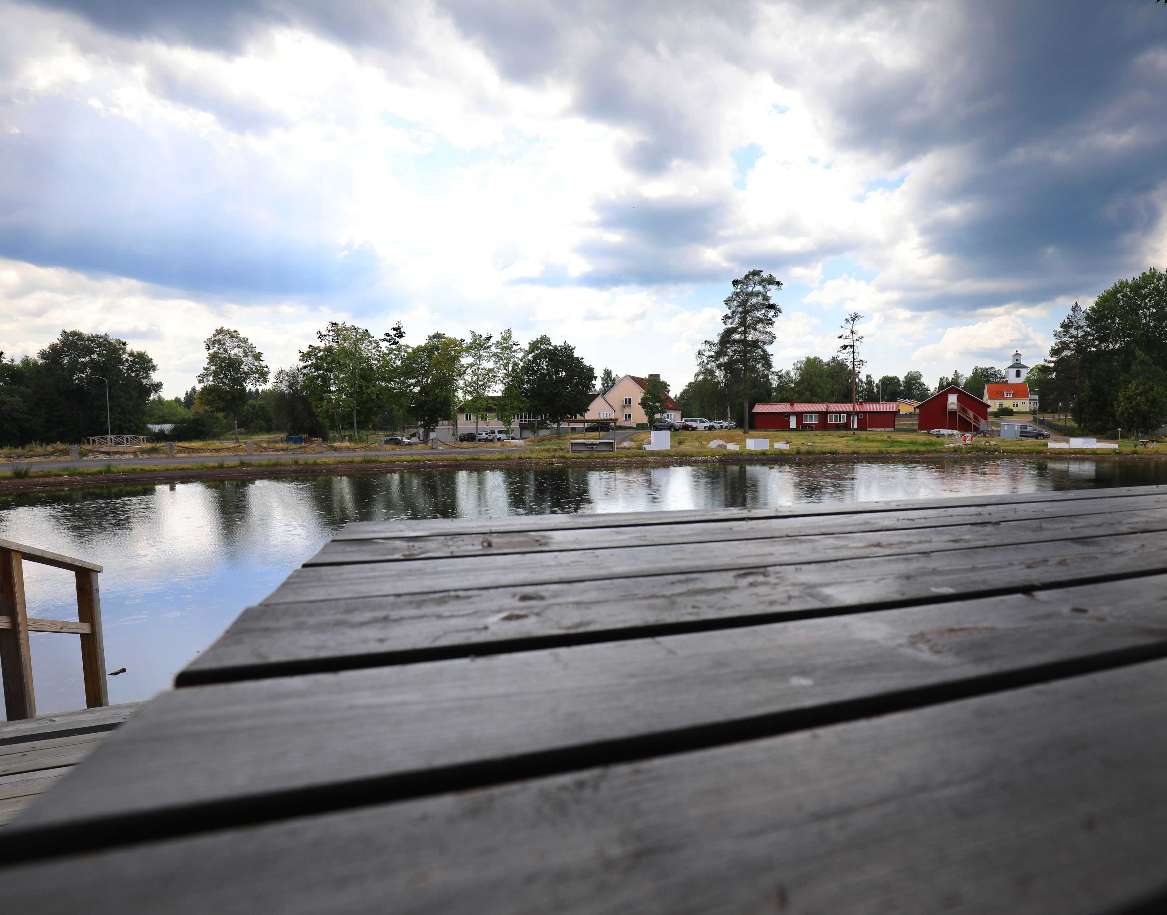 A wooden dock extending over a calm lake with a reflection of the sky and surrounding buildings.