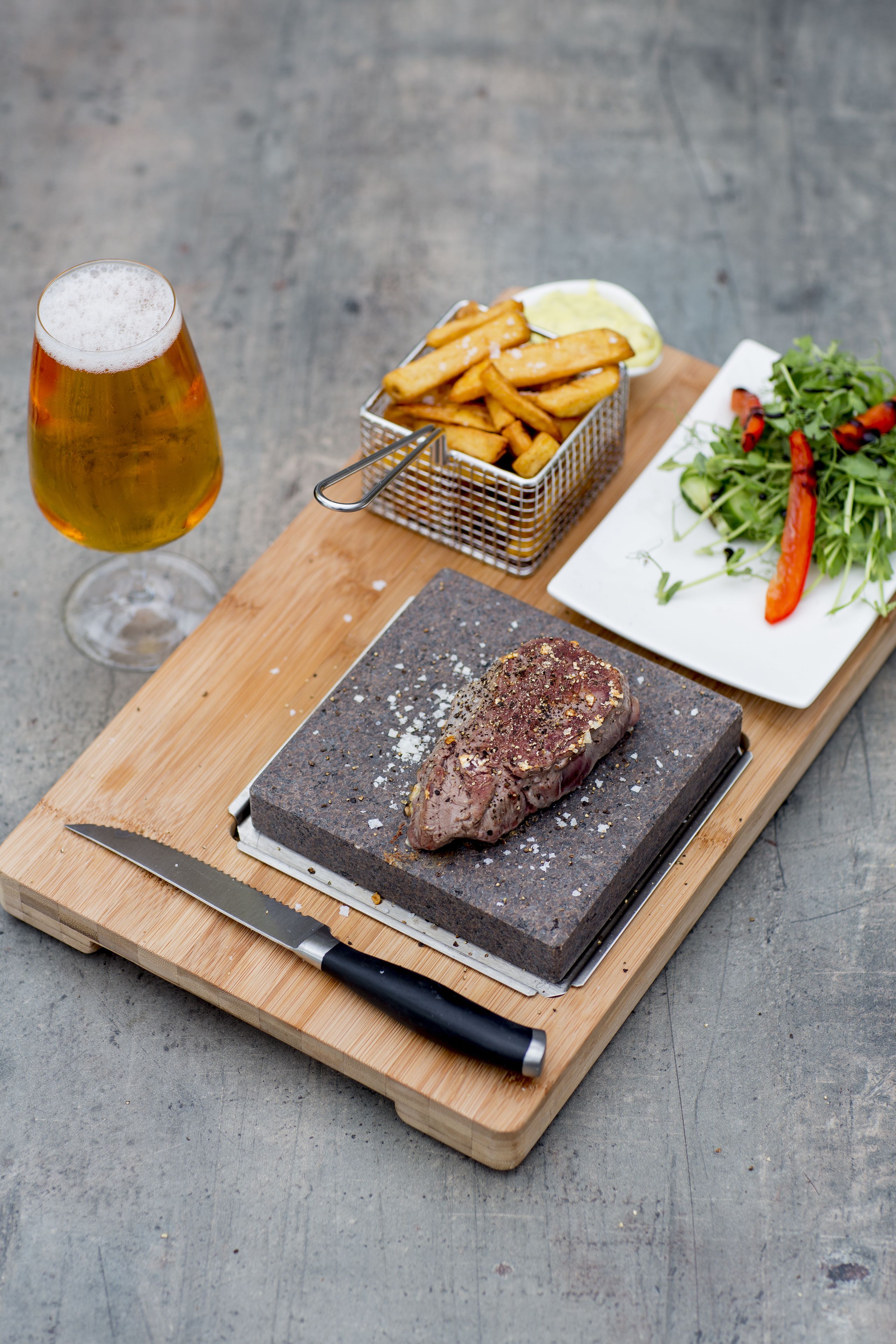 A steak on a cutting board with a knife, a glass of beer, a basket of fries, and a plate of greens.