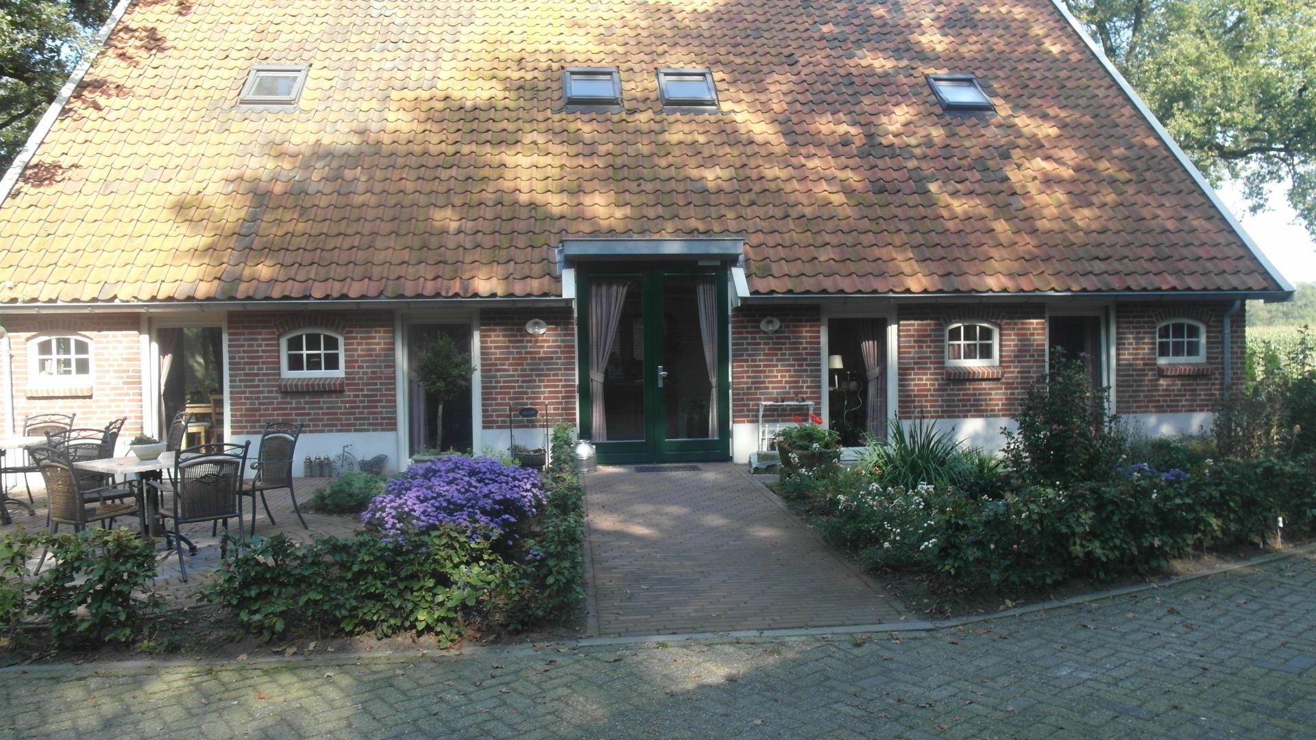 Ein zweistöckiges Backsteinhhaus mit Ziegeldach, einer grünen Tür und einer kleinen Veranda mit Gartenmöbeln.