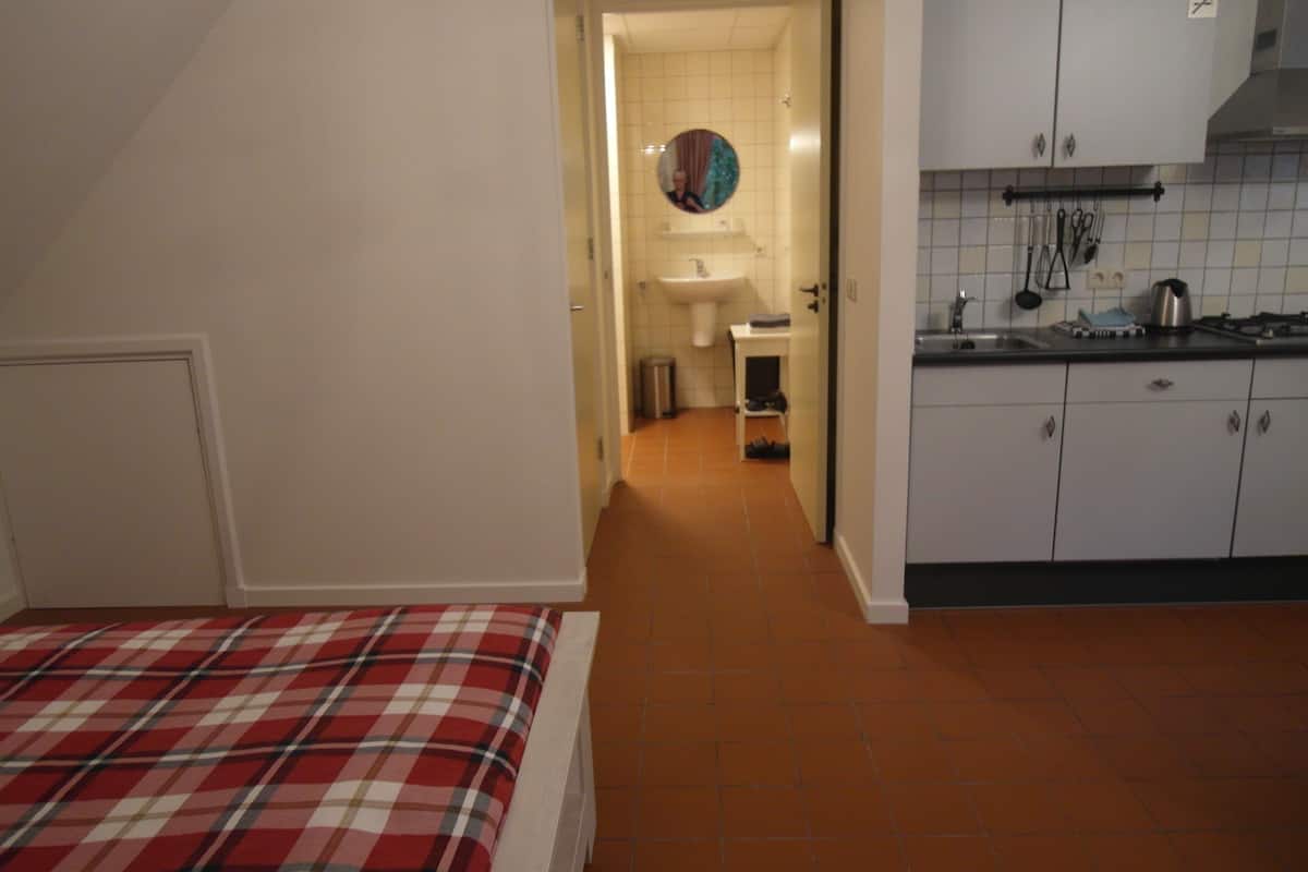Ein Schlafzimmer mit kariertem Überwurf, eine Küche mit weißen Schränken und einer gefliesten Spritzwand sowie ein Flur, der zum Badezimmer führt.