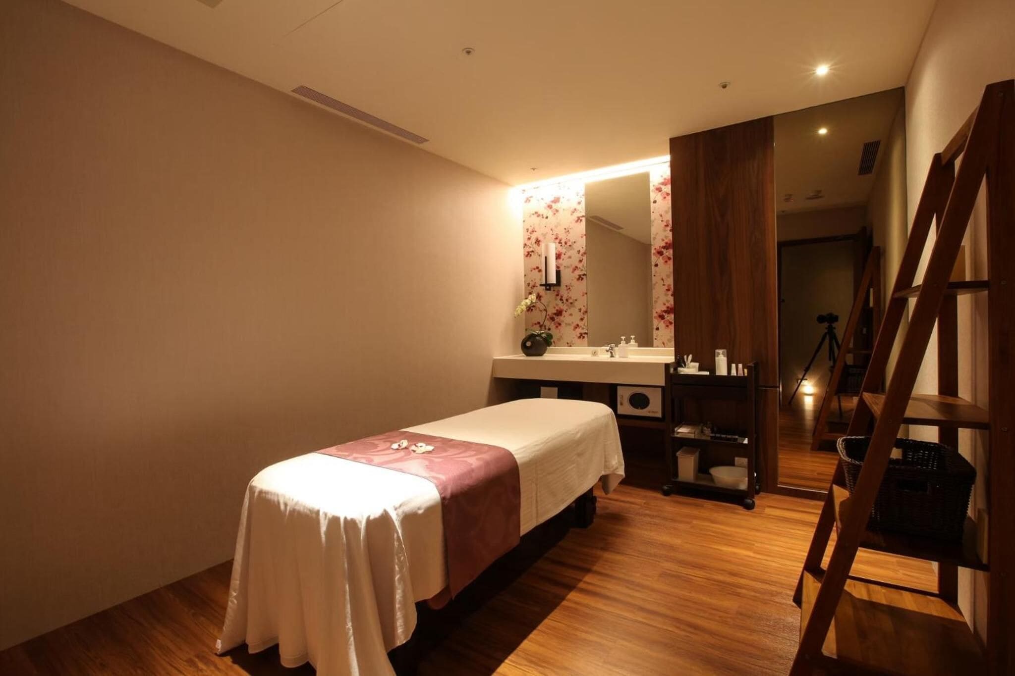 A single bed with a white and pink bedspread, a wooden floor, a wooden chair, a small table with a basket, and a mirror with floral wallpaper.