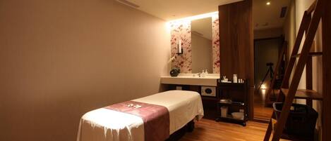 A single bed with a white and pink bedspread, a wooden floor, a wooden chair, a small table with a basket, and a mirror with floral wallpaper.