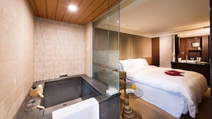 A modern hotel room with a large bed, a glass-enclosed shower, and a small sitting area with a table and chair.