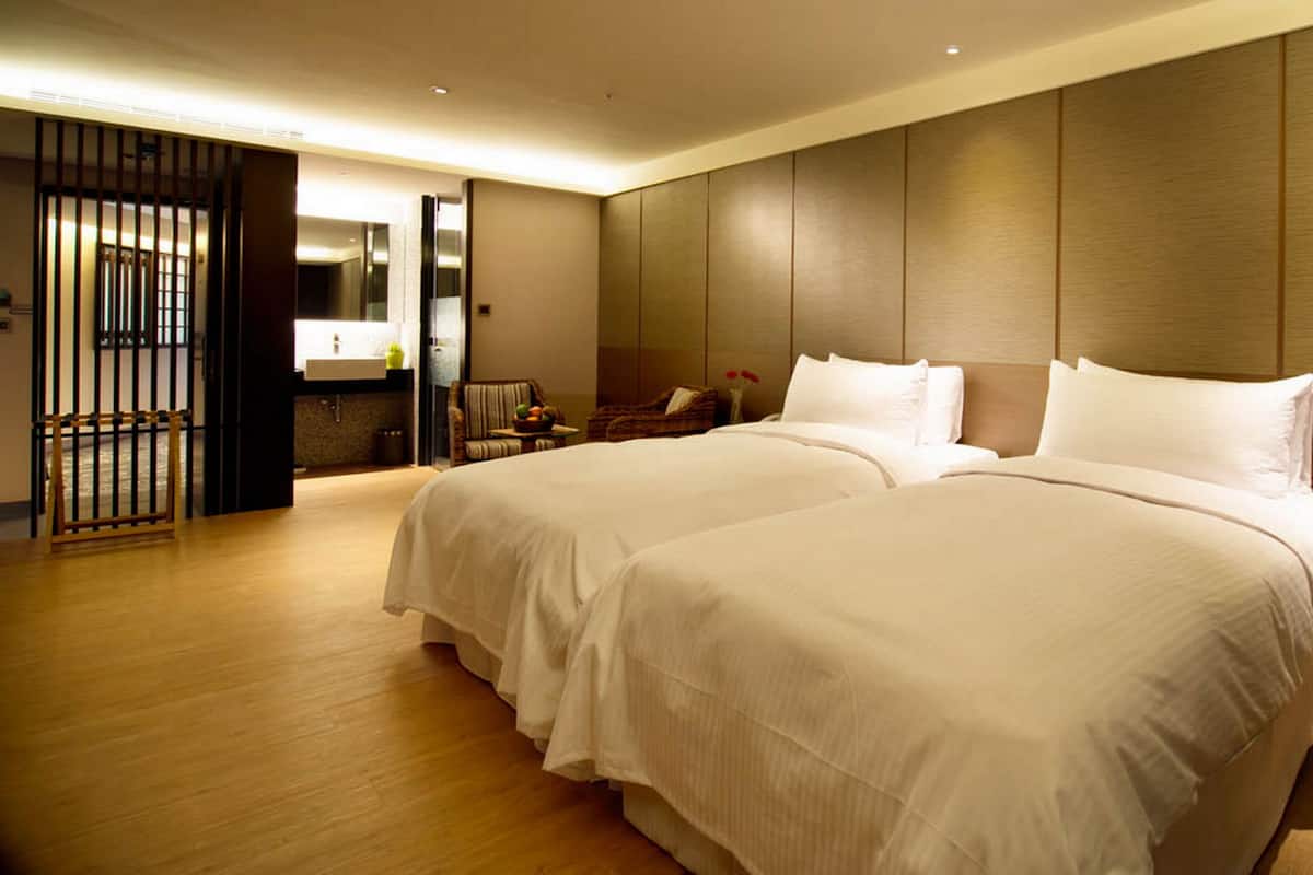 A hotel room with two large beds, a wooden floor, a modern bathroom, and a view of the outside.