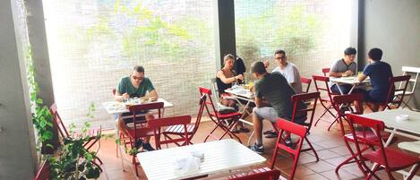 An outdoor dining area with red chairs and white tables, surrounded by a brick wall and a view of greenery.