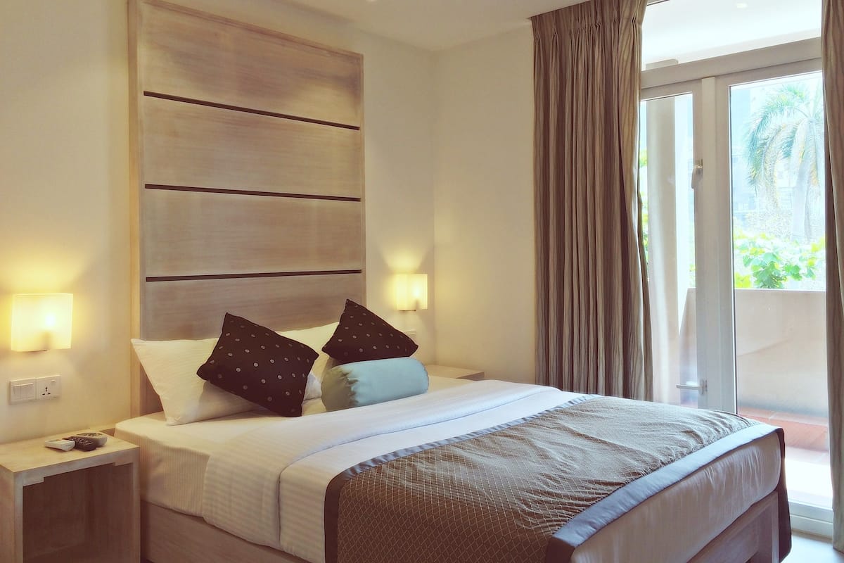 A hotel room with a large bed, two bedside tables, a wall-mounted headboard, and a view of the outdoors through a sliding glass door.