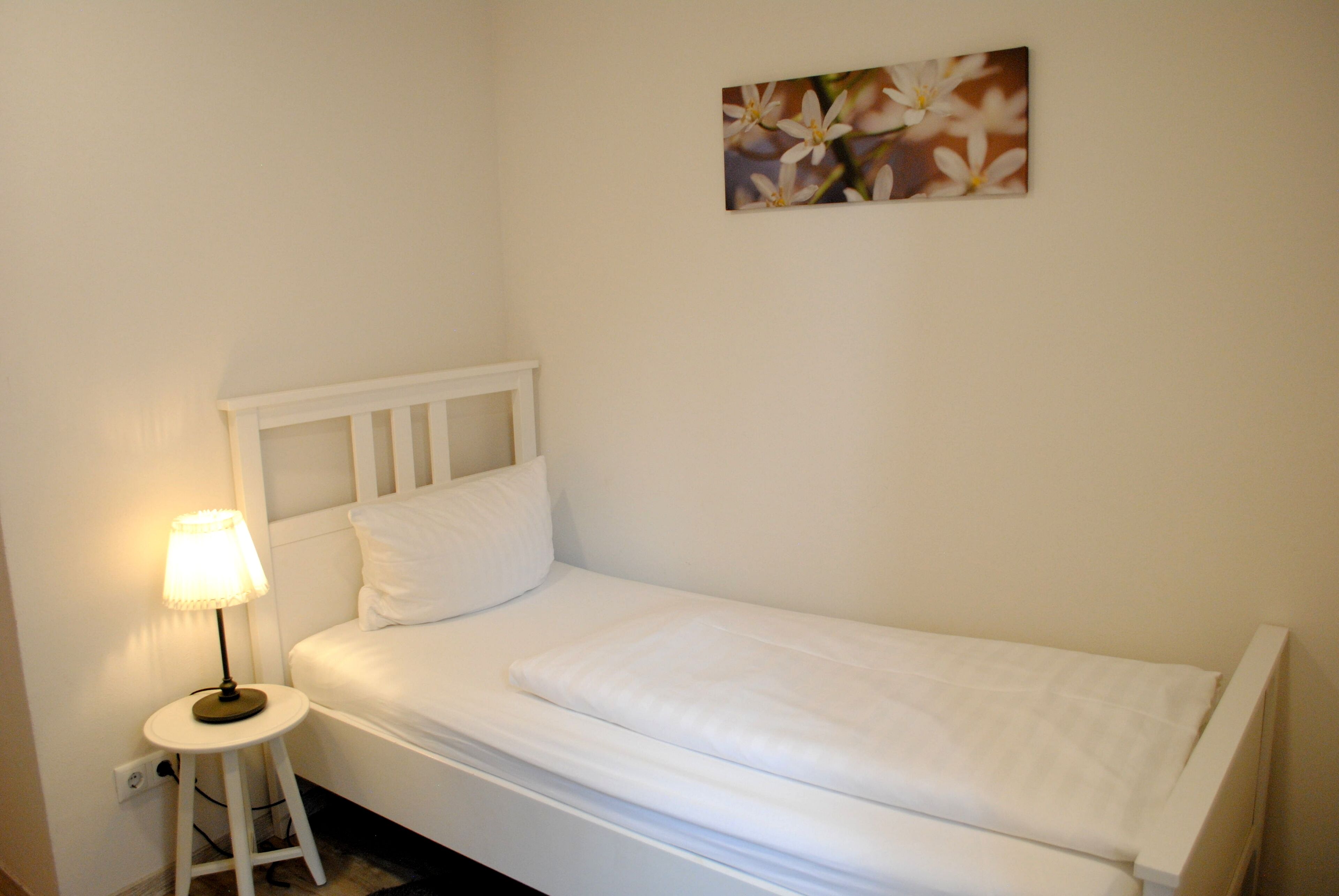 Una cama pequeña y bien arreglada con ropa blanca, un cabecero blanco y una lámpara de noche.