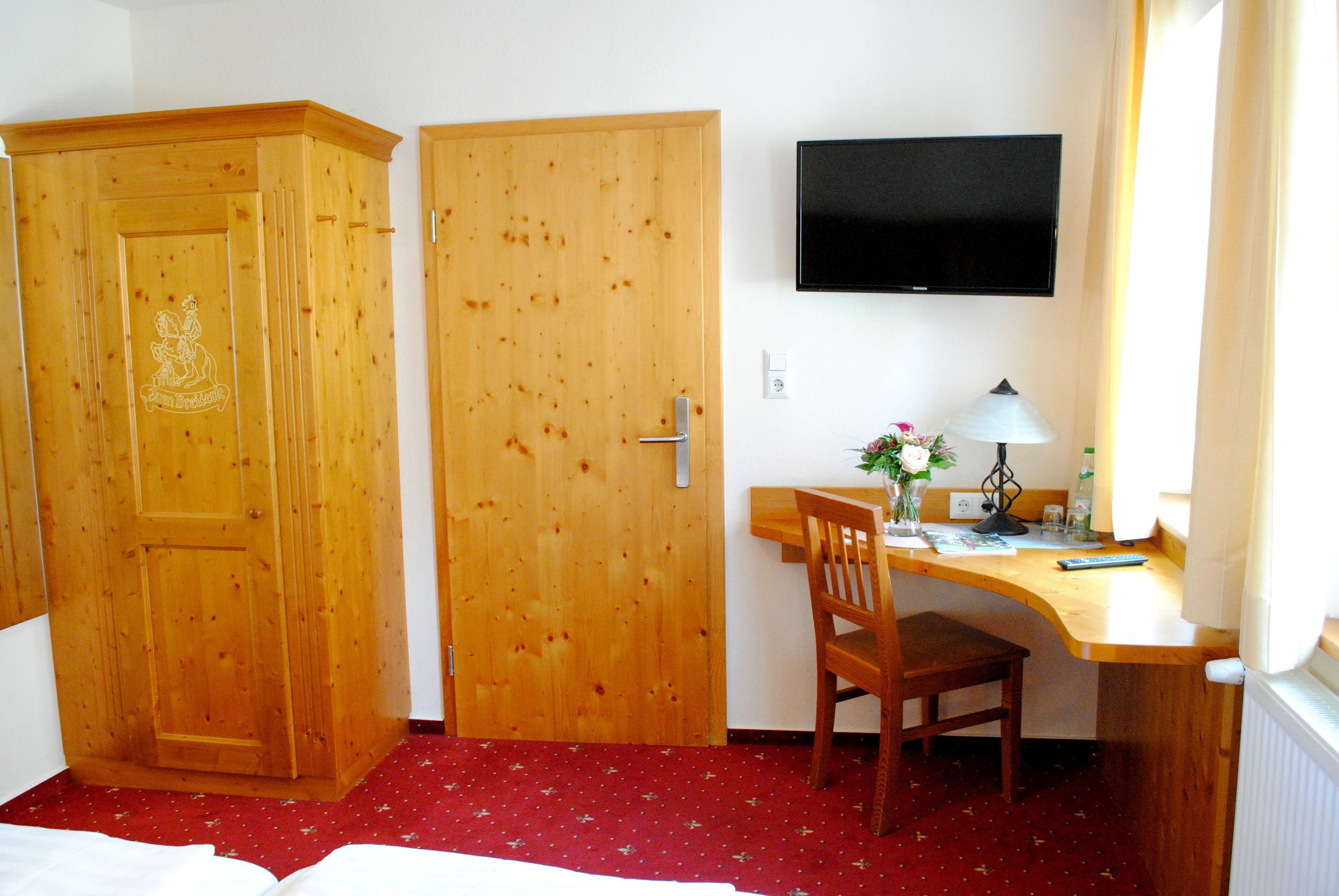 A room with a wooden wardrobe, a desk with a chair, a television mounted on the wall, and a window with curtains.