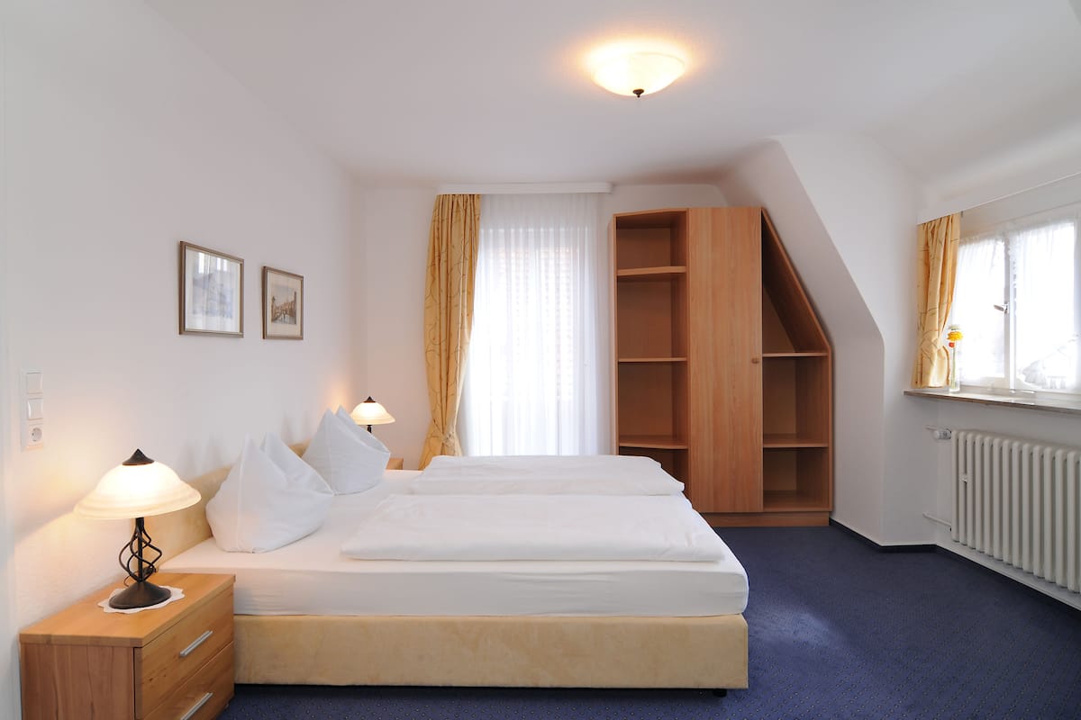 Una habitación de hotel con cama, lámparas de noche, un armario de madera y una ventana con cortinas.