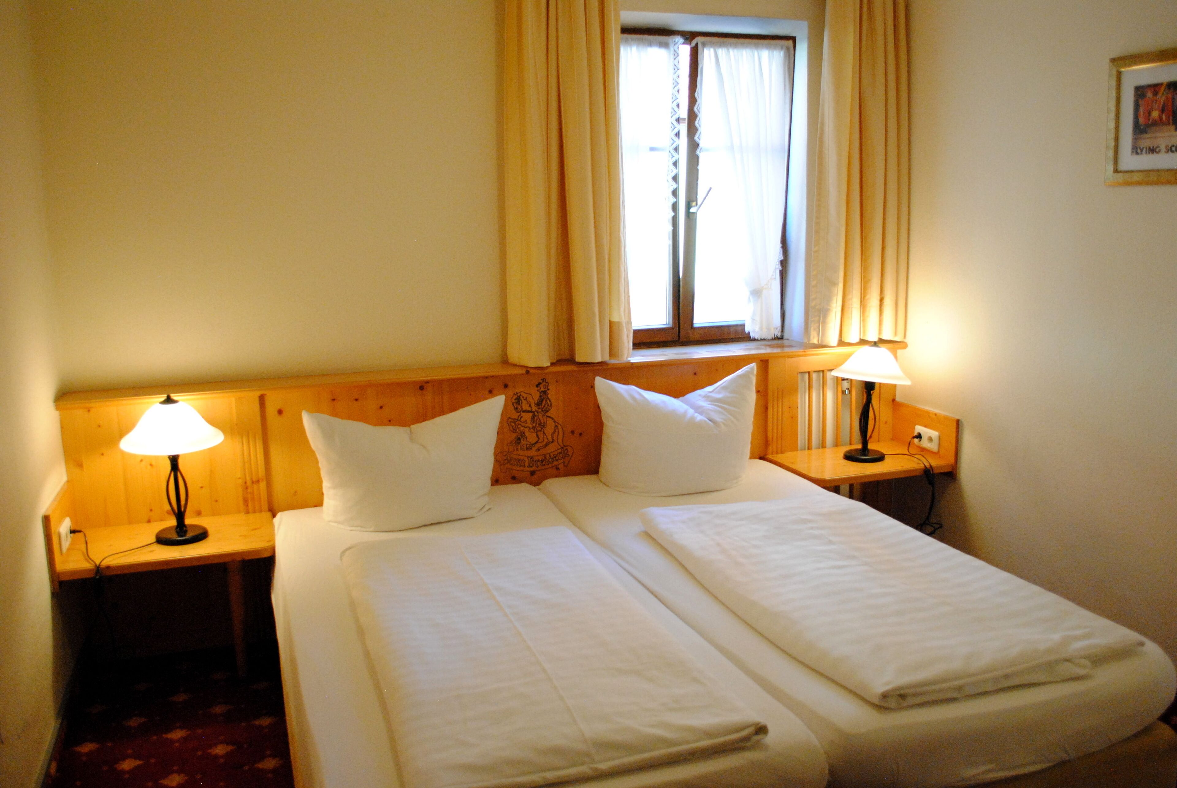 A double bed with white linens, two bedside tables with lamps, a window with curtains, and a wooden headboard.