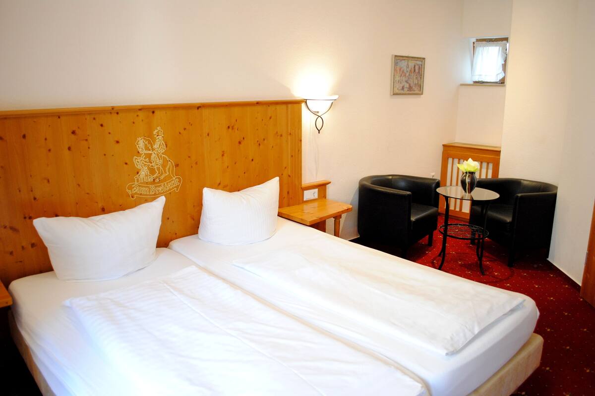 Habitación de hotel con dos camas individuales, cabecera de madera, mesita de noche, una pequeña mesa, dos sillas negras, alfombra roja y una lámpara de pared.