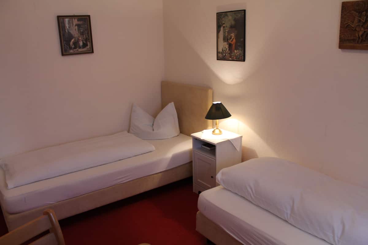 Una habitación de hotel pequeña y con una cama individual, una mesita de noche con lámpara y dos cuadros enmarcados en la pared.