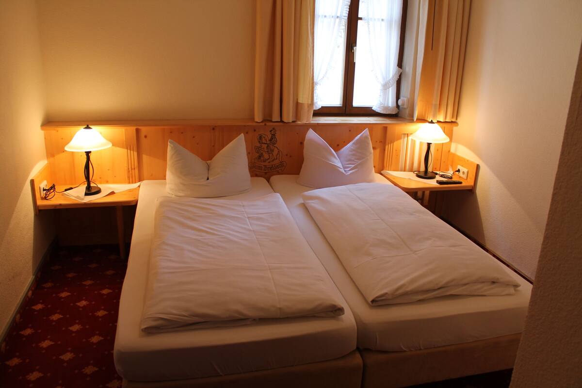 Una cama doble con dos mesitas de noche, cada una con una lámpara, en una habitación con cabecera de madera y ventana con cortinas.