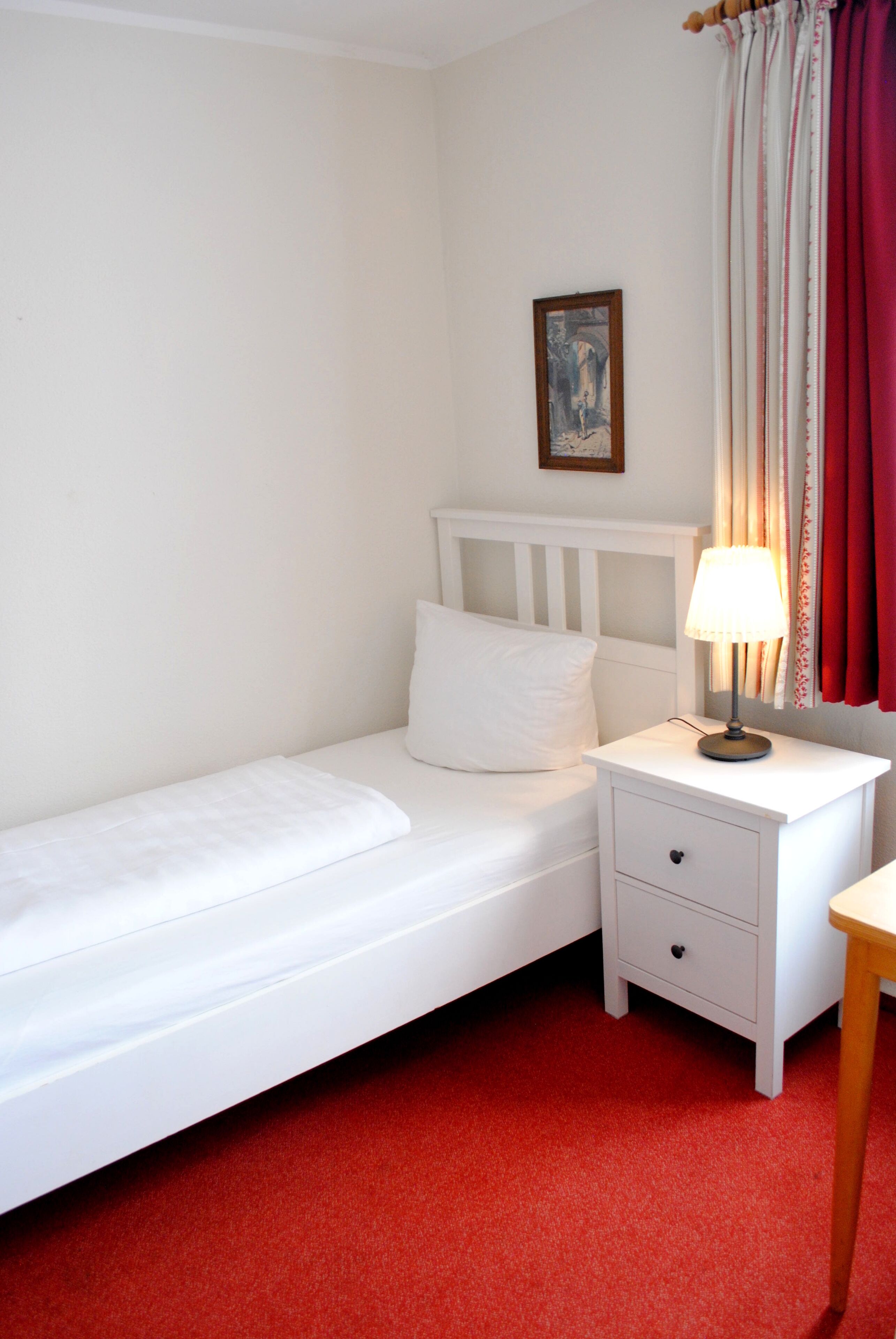 Una cama blanca con una mesita de noche, una lámpara y un pequeño cuadro en la pared.