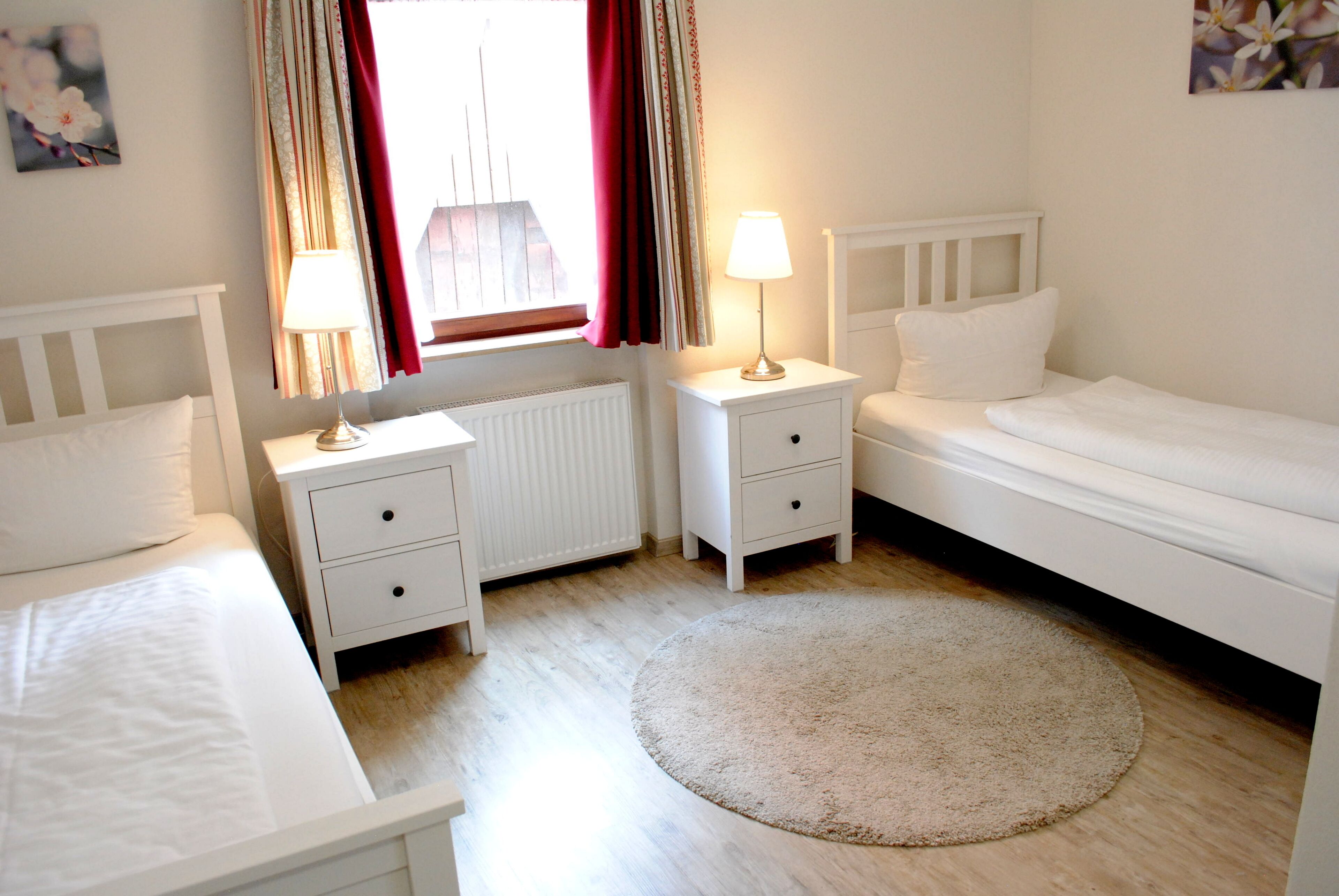 A room with two single beds, a round rug, a window with curtains, and a radiator.
