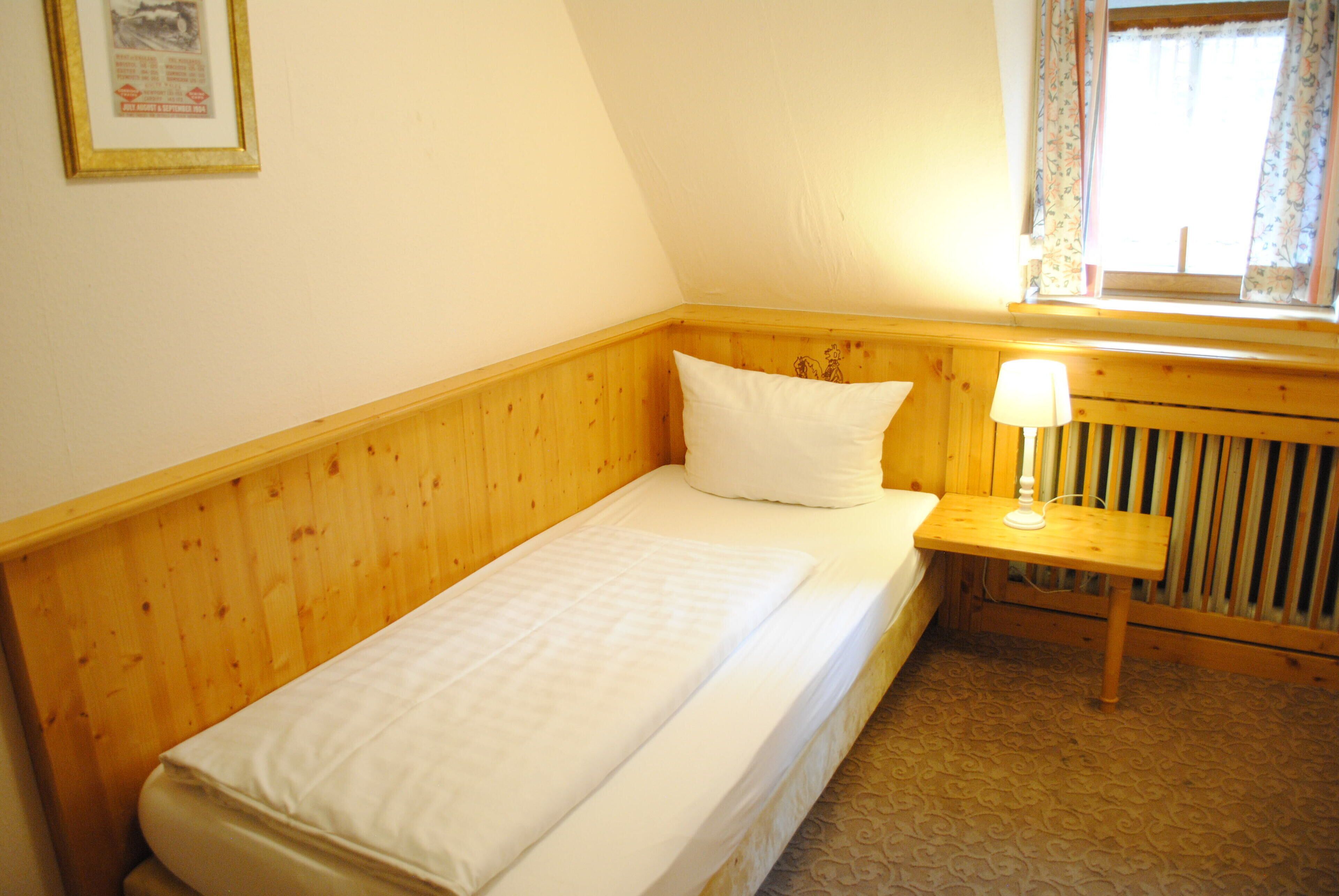 A small, single-bed room with wooden paneling, a bedside table with a lamp, and a radiator.