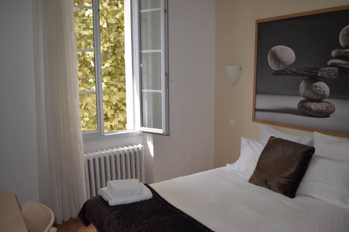 A bedroom with a bed, a window with curtains, a radiator, a painting on the wall, and a chair.