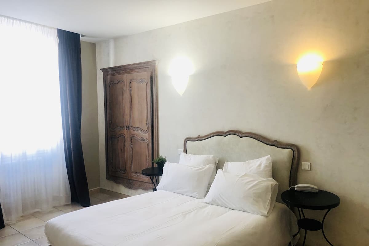 A hotel room with a bed, a nightstand, a wall-mounted light, a wardrobe, and a window with curtains.