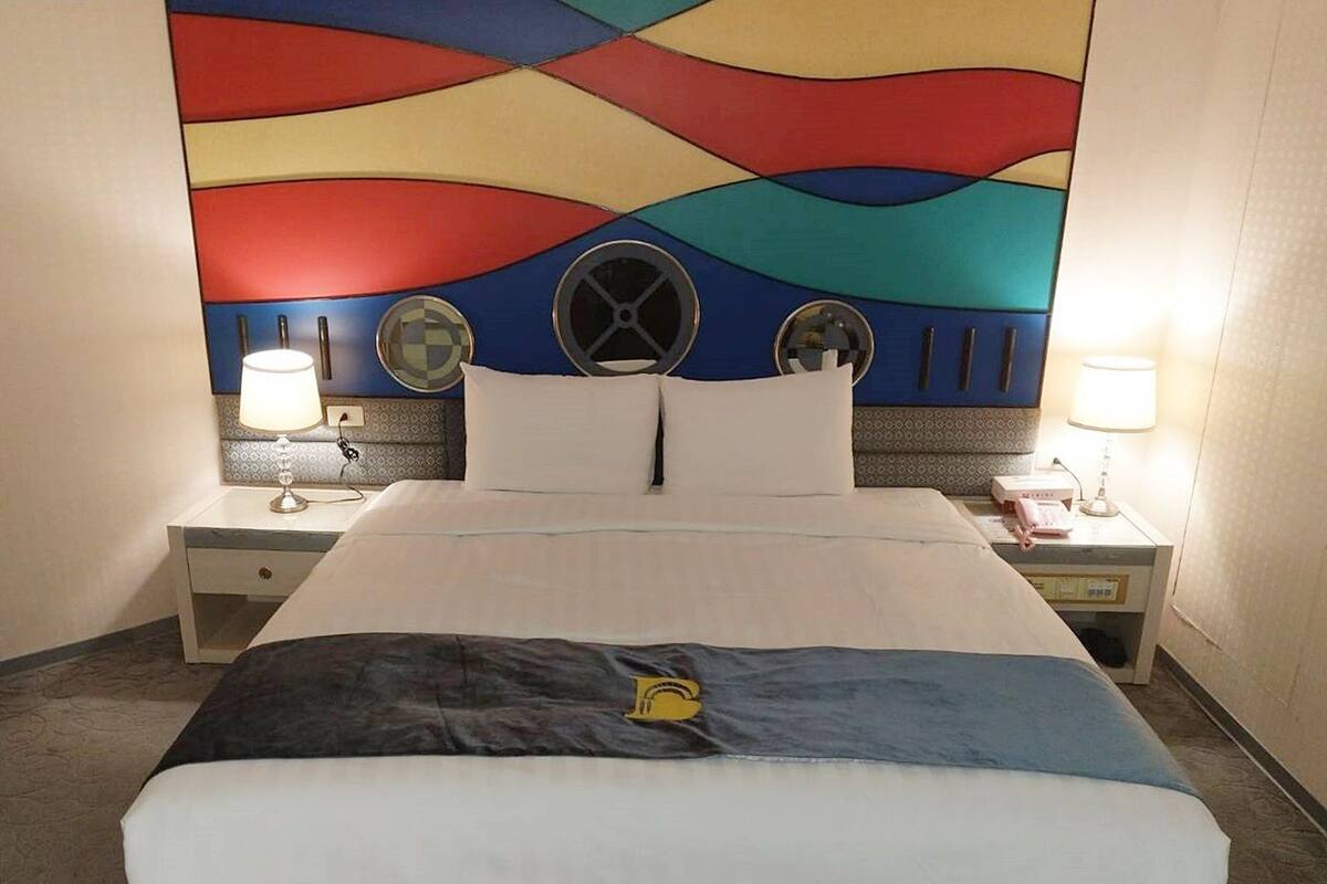 Habitación de hotel con una cama grande, dos mesitas de noche, un mural abstracto y colorido en la pared, y dos ventiladores de pared.