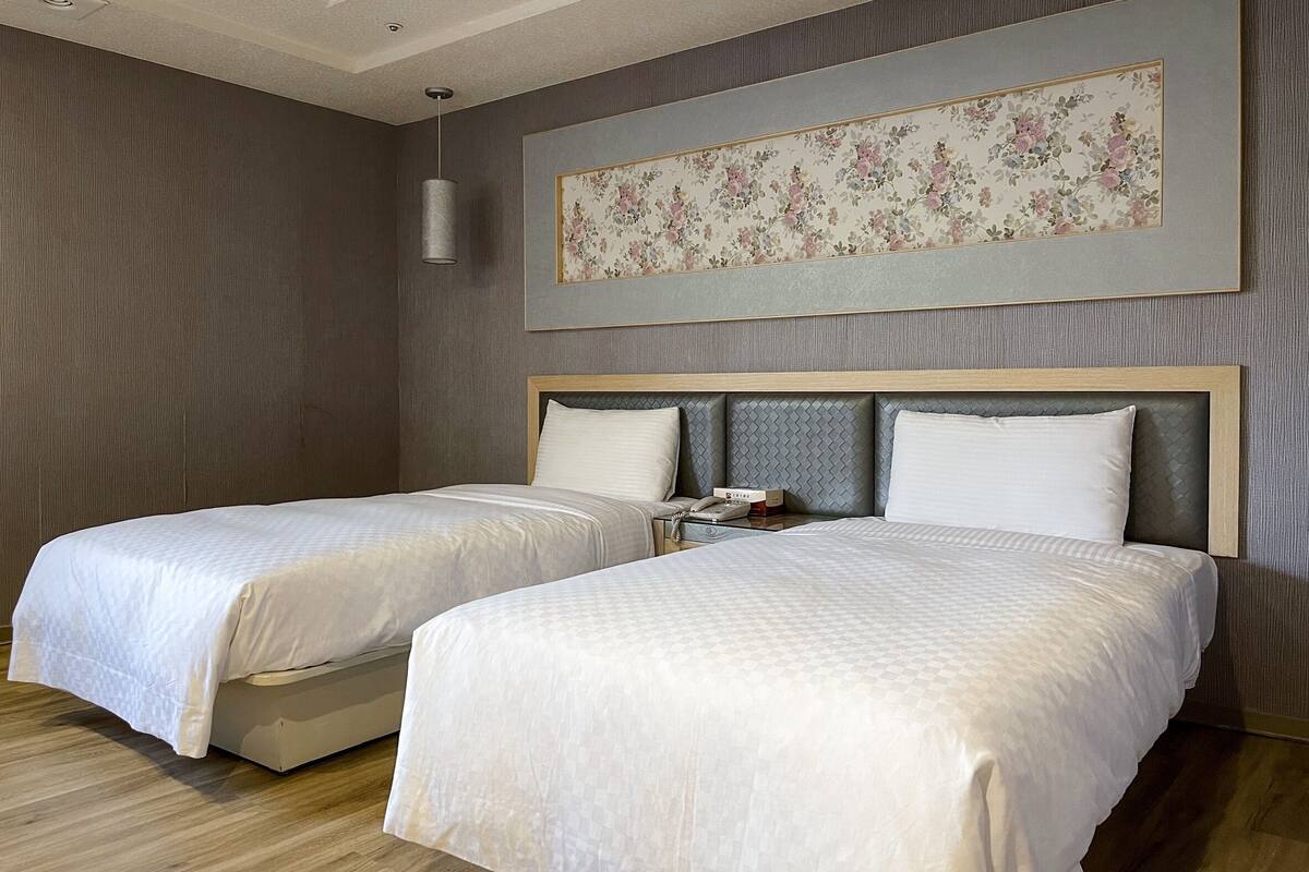 Habitación de hotel con dos camas, cabecero con diseño texturizado, cuadro floral enmarcado en la pared y mesita de noche con un teléfono y una caja.