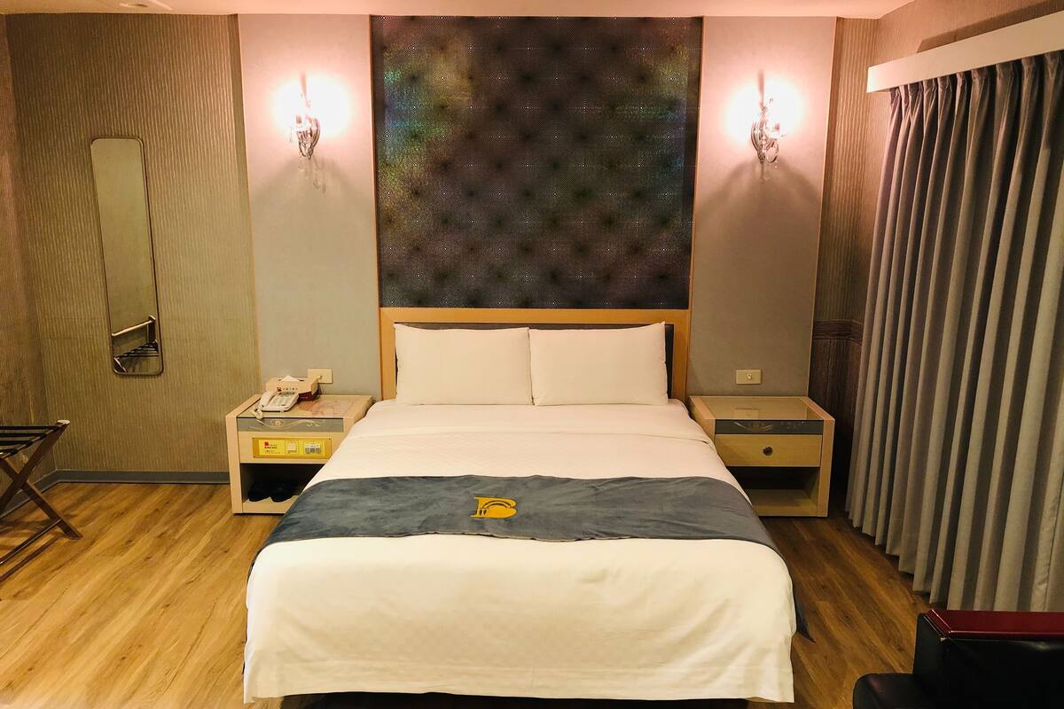 Habitación de hotel con una cama grande, dos mesitas de noche, un espejo, una obra de arte en la pared y una cortina.