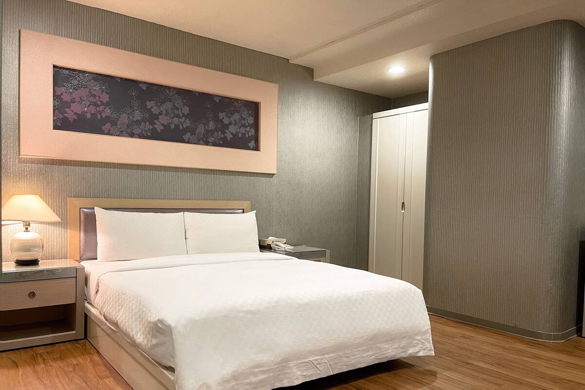 Habitación de hotel ordenada con cama, mesita de noche, lámpara y un cuadro grande con motivos florales en la pared.