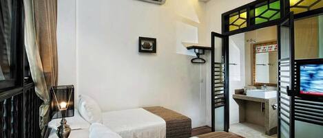 A hotel room with two beds, a bedside table with a lamp, an air conditioning unit, a bathroom with a sink and mirror, and a television.