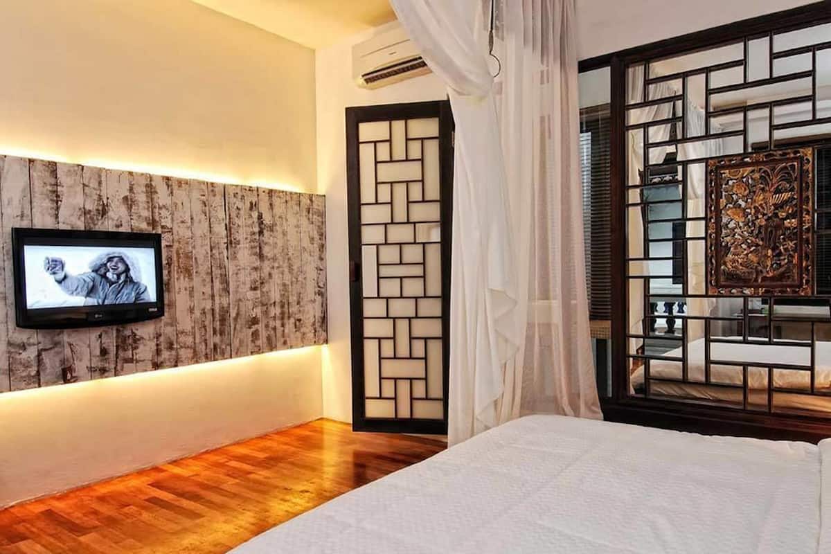 A bedroom with a wooden floor, a bed with white bedding, a television mounted on a wooden wall, a traditional decorative screen, and a window with curtains.